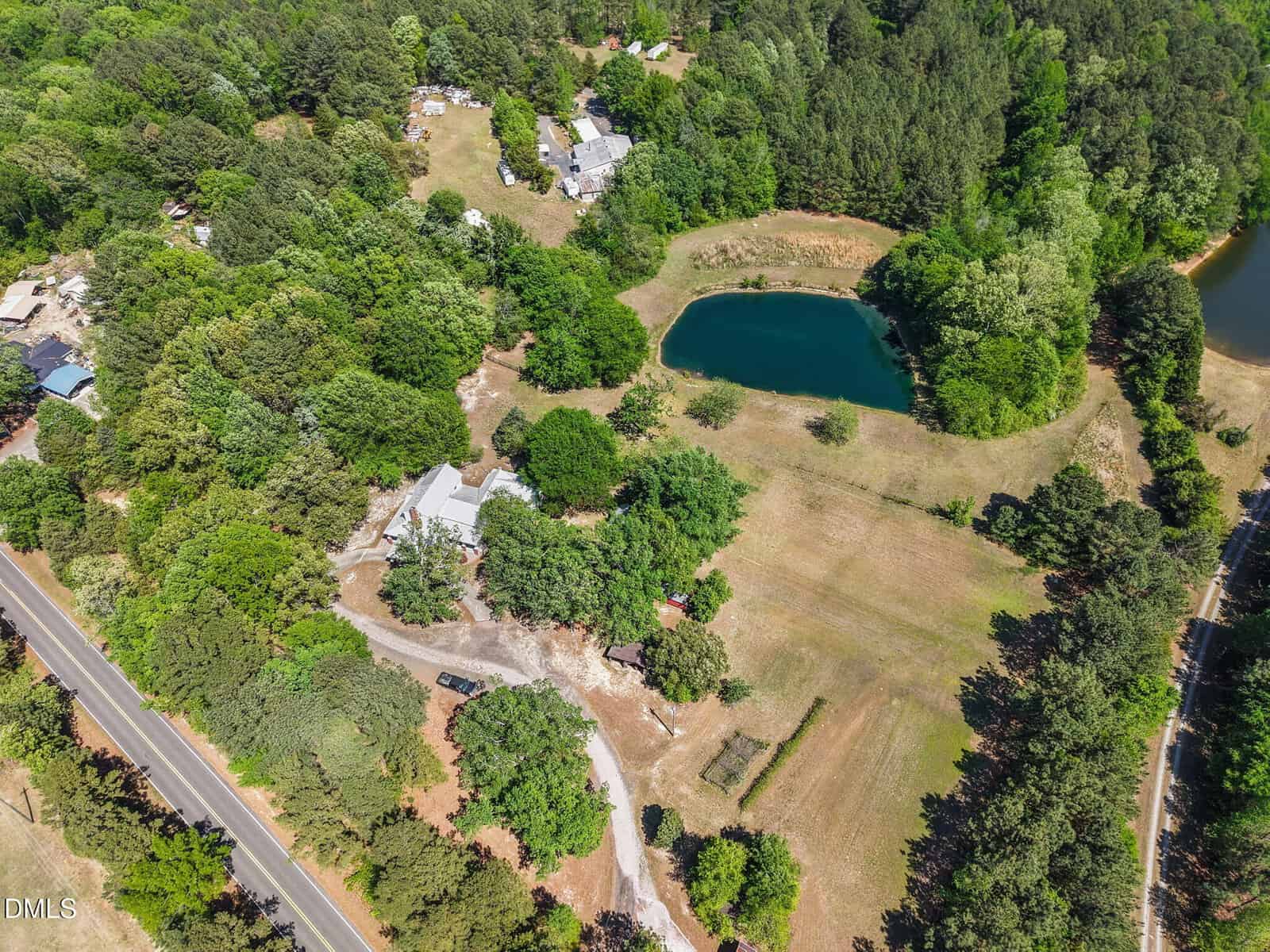 Aerial view of a rural property with a pond, surrounded by trees and grass fields. Several buildings and houses are scattered across the landscape, with a road running along the edge of the property.