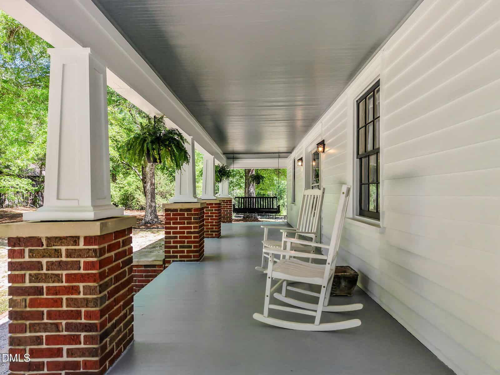 A spacious covered porch with white siding, two white rocking chairs, brick columns, hanging plants, and a dark porch swing at the far end, overlooking a green yard with trees.