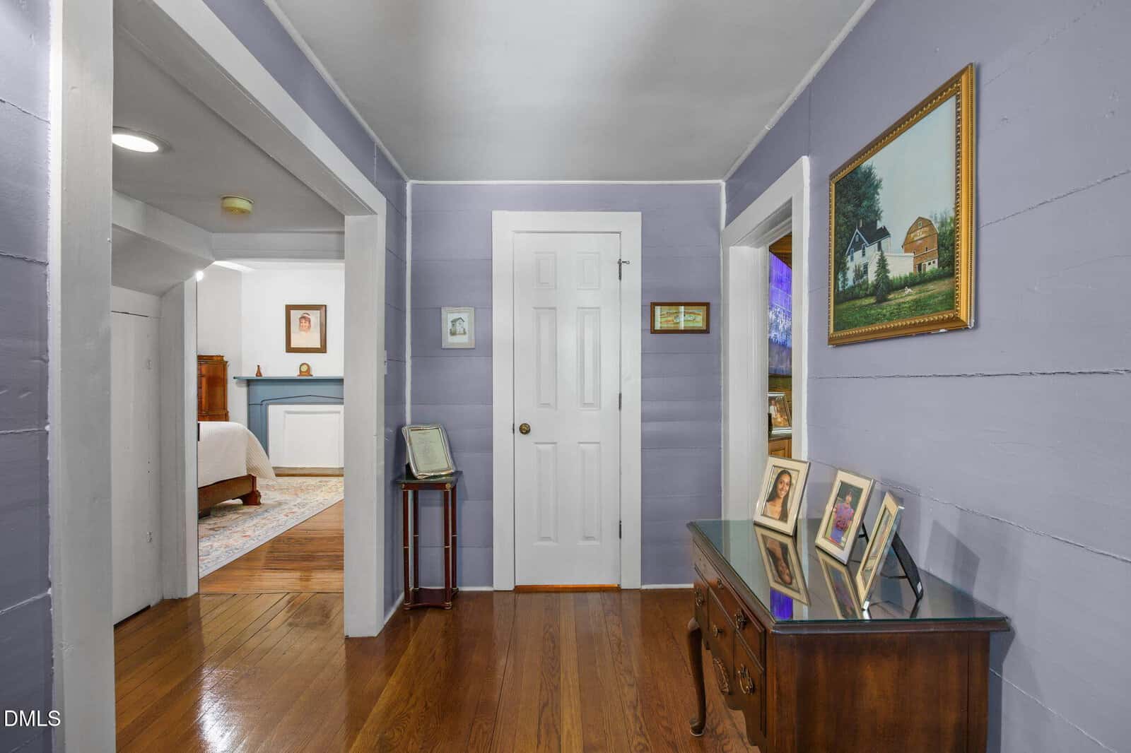 A hallway with lavender walls, hardwood floors, family photos on a glass-topped wooden table, framed art on the walls, and doorways leading to other rooms including a bedroom with a blue and white color scheme.