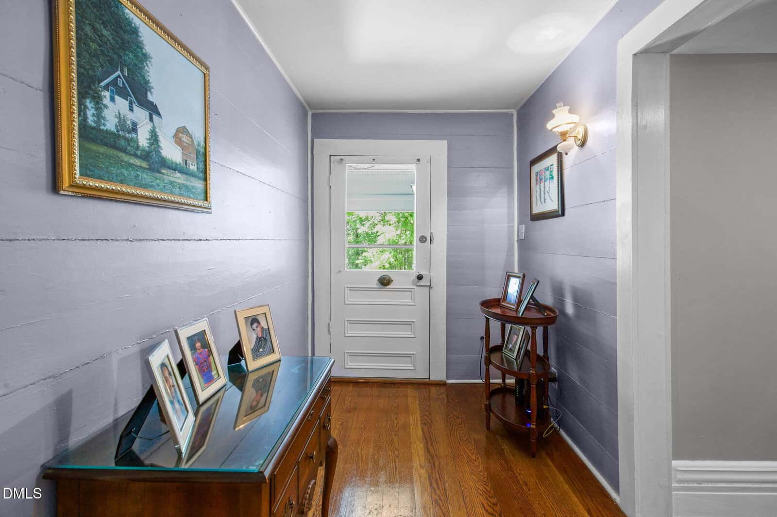A hallway with lavender walls, wooden floor, a white door at the end, framed photos on a chest and small table, a wall sconce, and paintings hanging on the walls.