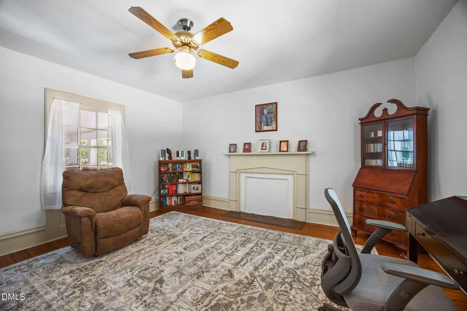 Bright room with a ceiling fan, brown recliner, bookshelf, white fireplace, glass-front cabinet, desk chair, and large patterned rug. Window with sheer curtain lets in natural light.