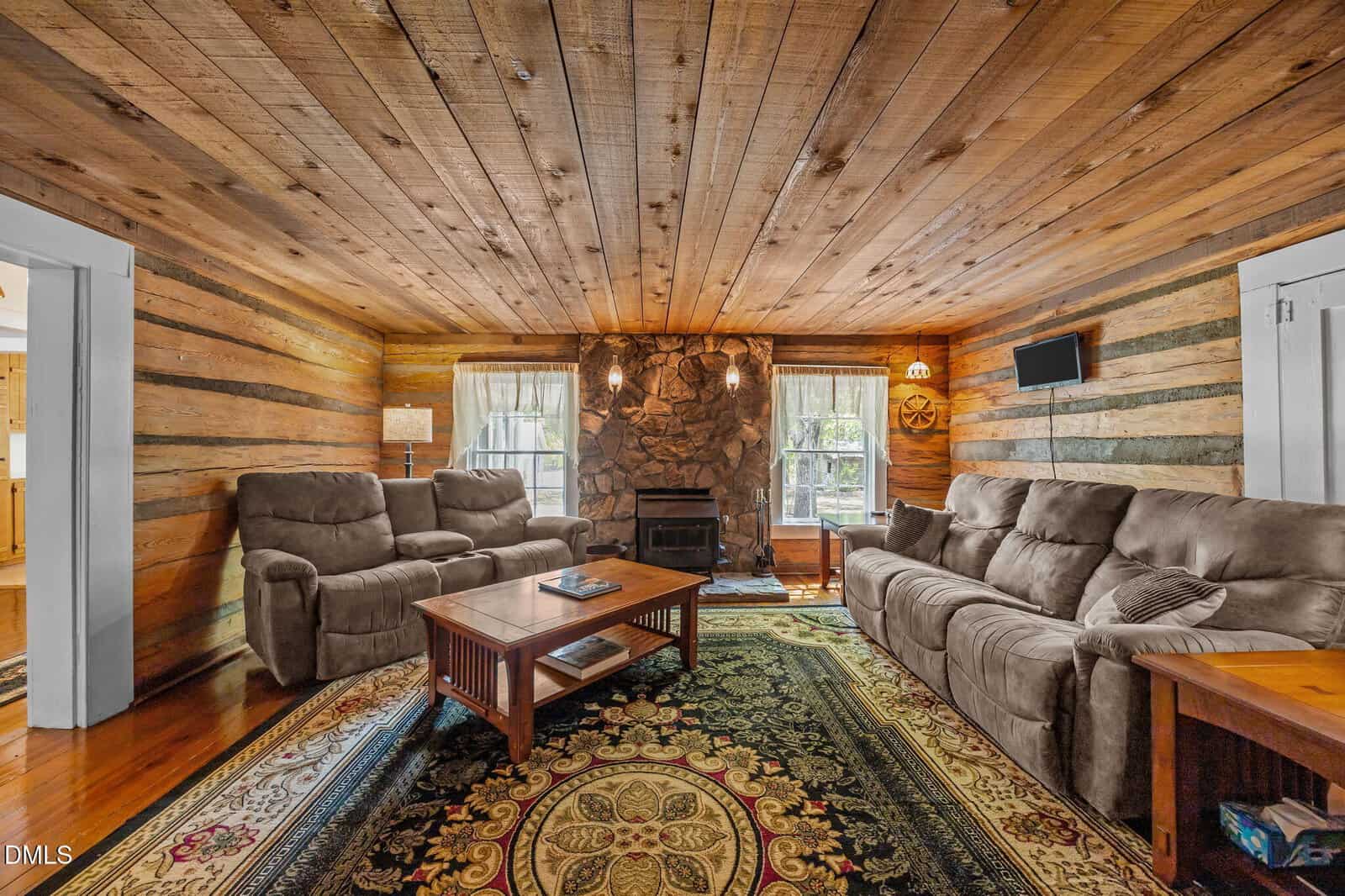Cozy log cabin living room with rustic wood walls and ceiling, stone fireplace, beige sofas, a wooden coffee table, and a patterned area rug. Large windows let in natural light.