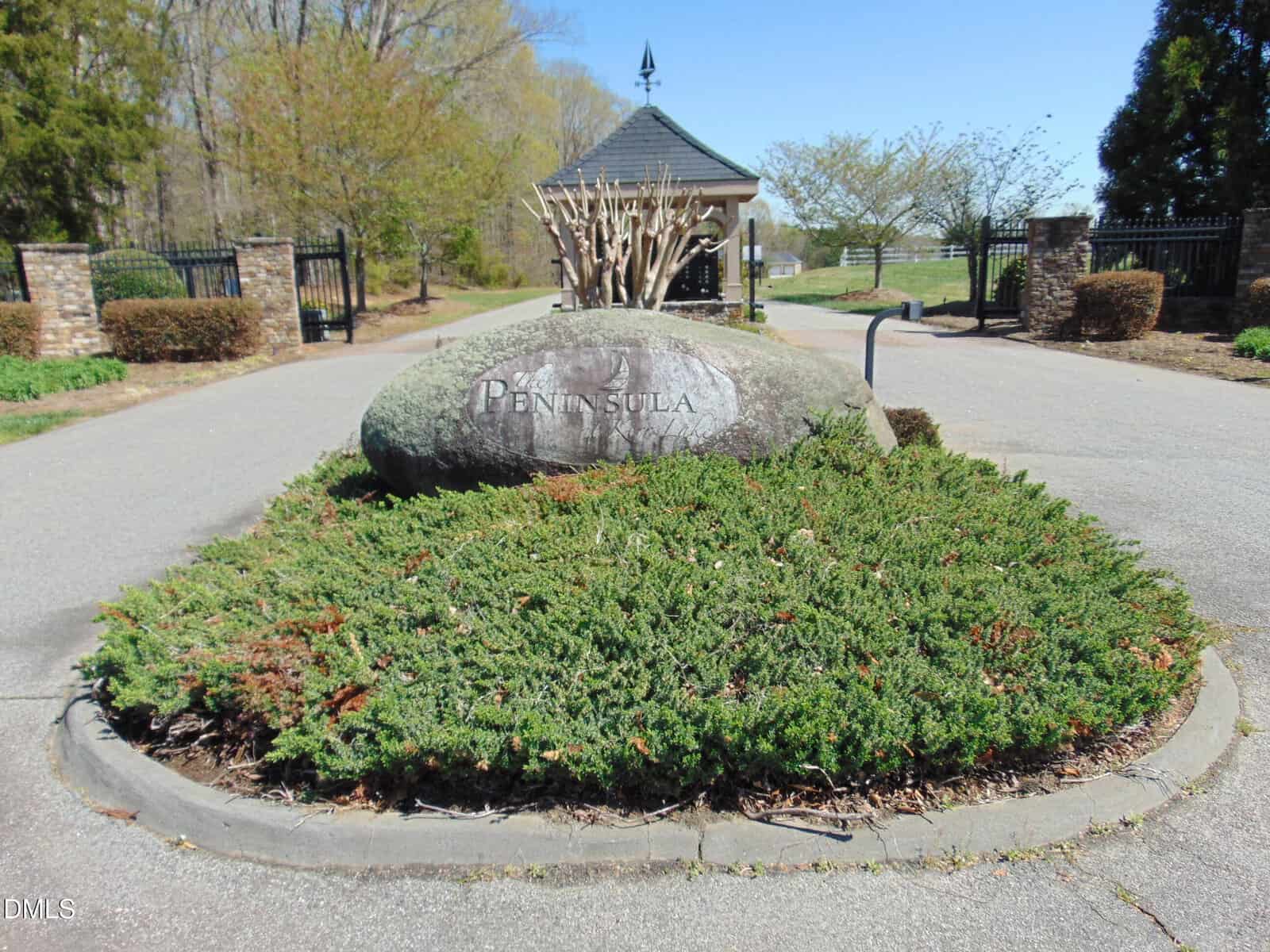 A large stone engraved with "PENINSULA" sits in the center of a landscaped roundabout at the entrance to a gated community, with brick pillars and black metal gates in the background.