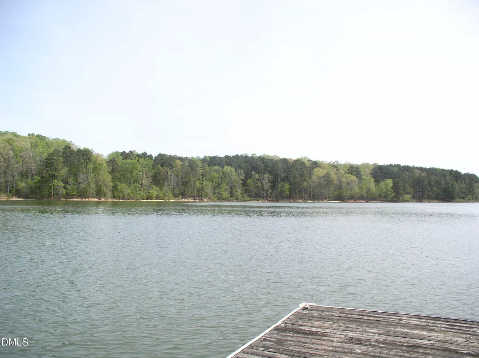 A calm lake with a forested shoreline in the background, viewed from the edge of a wooden dock on a clear, bright day.