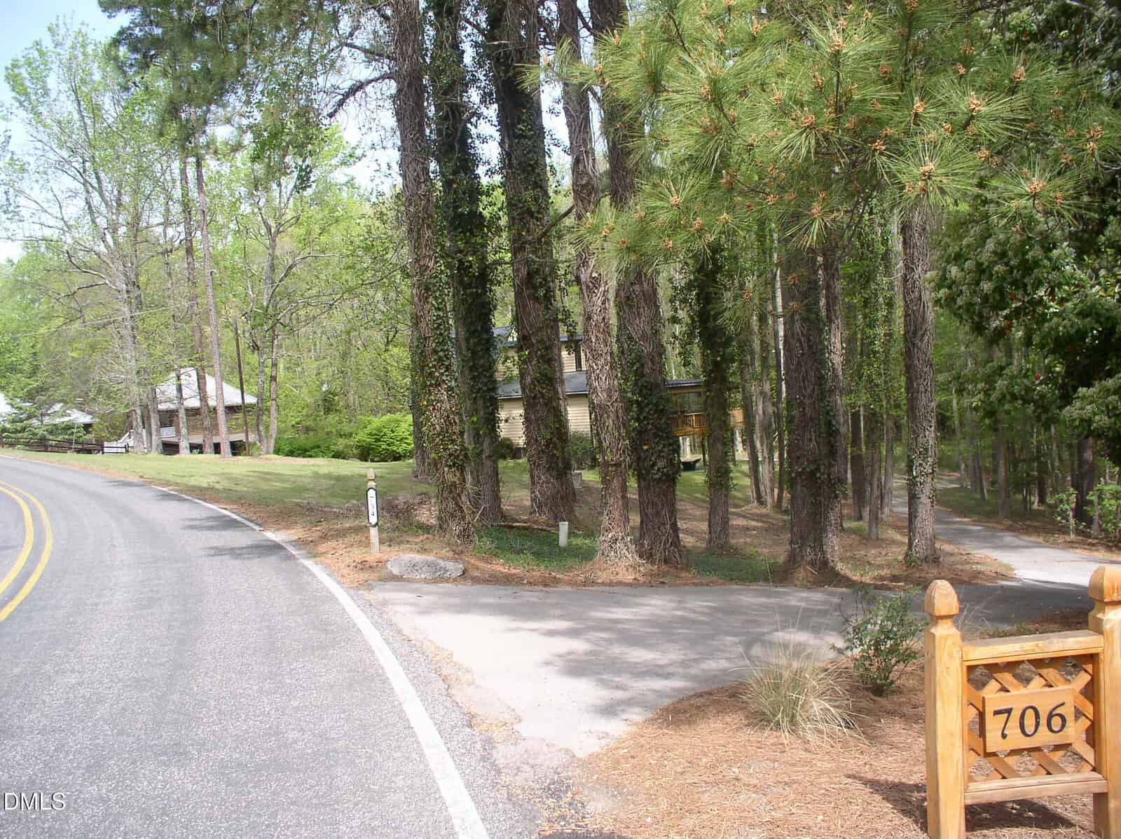 A winding paved road curves left beside a tall pine and leafy tree line. A wooden post marked "706" stands near a driveway leading into the trees, with a house visible in the background.