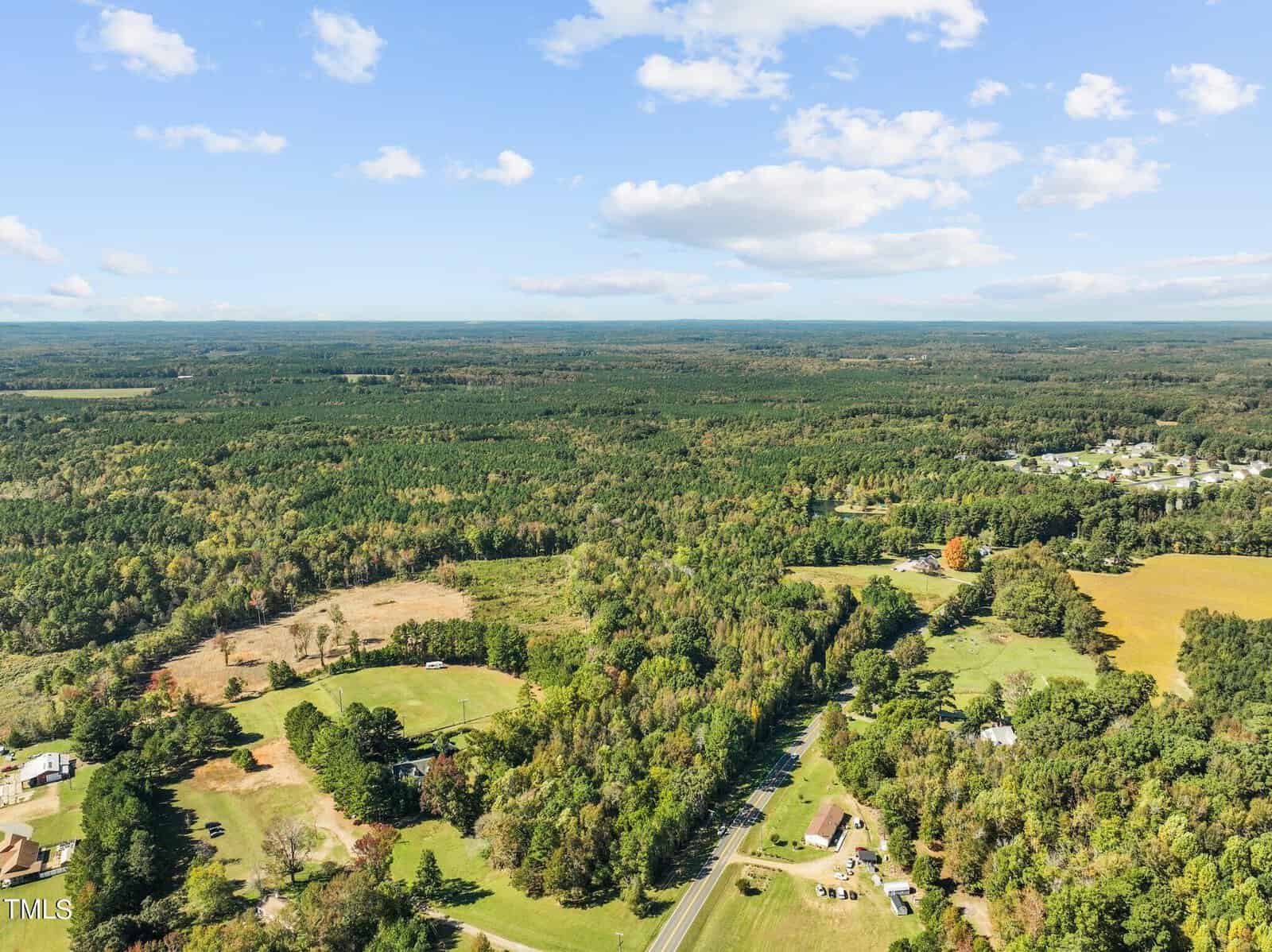 Aerial view of a rural landscape with expanses of green forest, scattered houses, fields, and a road winding through the area under a blue sky with scattered clouds.