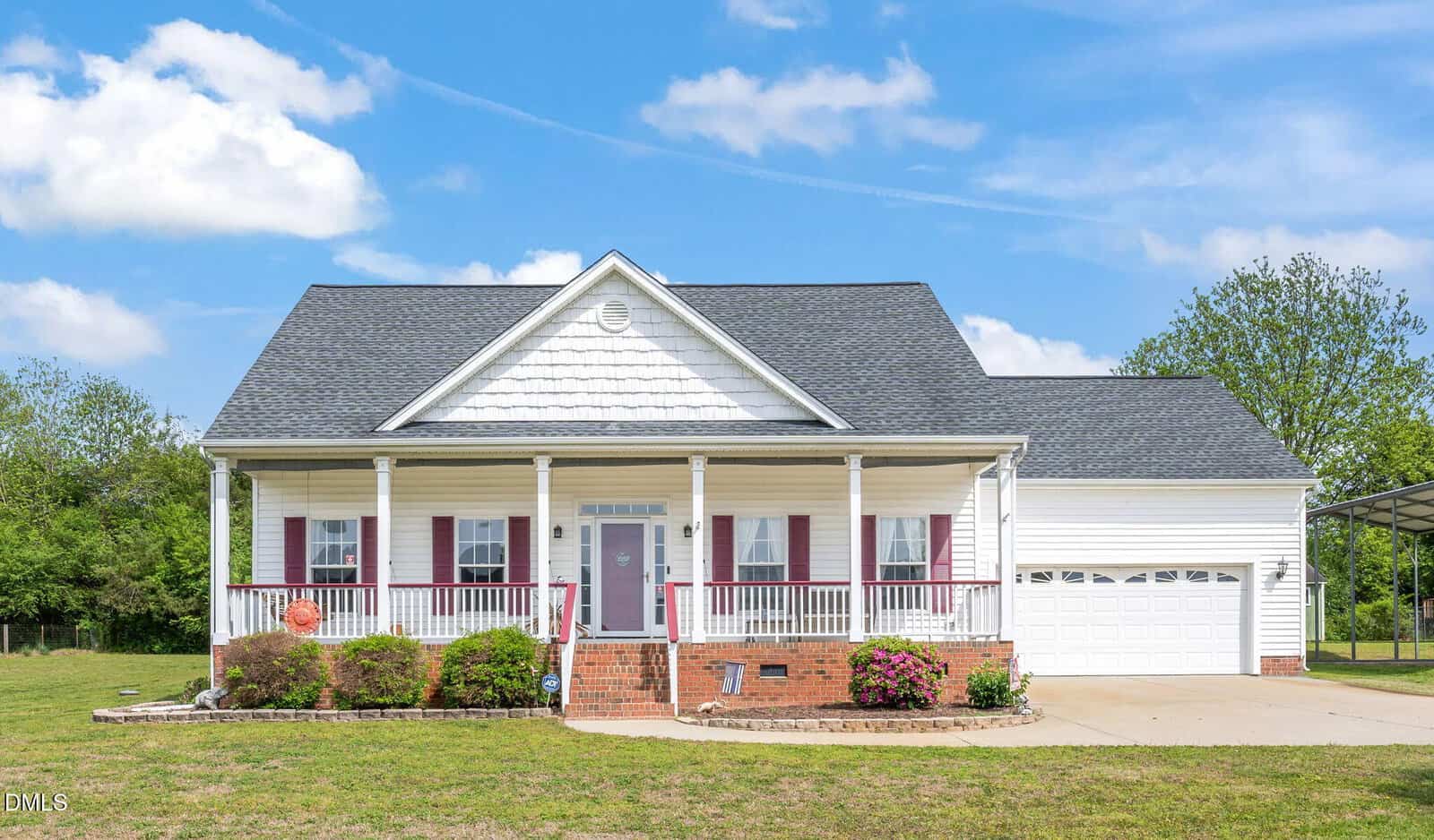 A white single-story house with a gable roof, red shutters, and a covered front porch. There is a two-car garage to the right, a brick walkway, flower beds, and a well-kept lawn under a blue sky with scattered clouds.