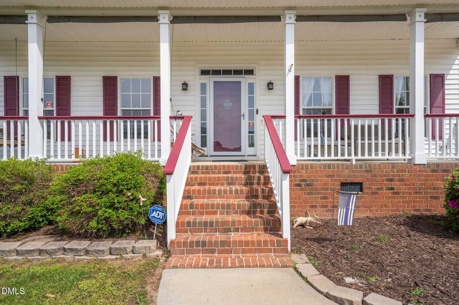 Front view of a house with a brick staircase leading to a red front door, white railing, and columns. Purple shutters frame the windows, and a security sign and American flag are in the landscaped yard.