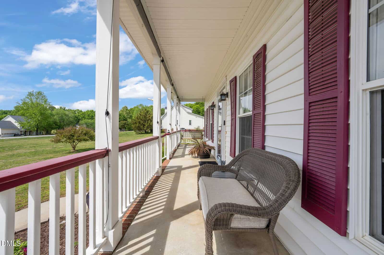 A spacious front porch with a white railing, maroon shutters, and a wicker bench overlooks a grassy yard and trees under a blue sky with scattered clouds.