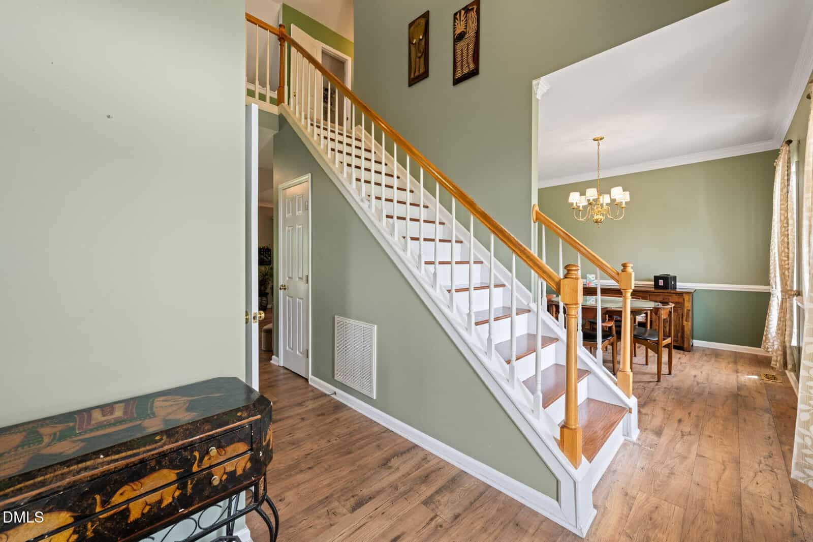 A staircase with white railings and wooden steps leads to an upper floor in a home with green walls and wooden flooring. To the right is a dining room with a chandelier and large windows with curtains.