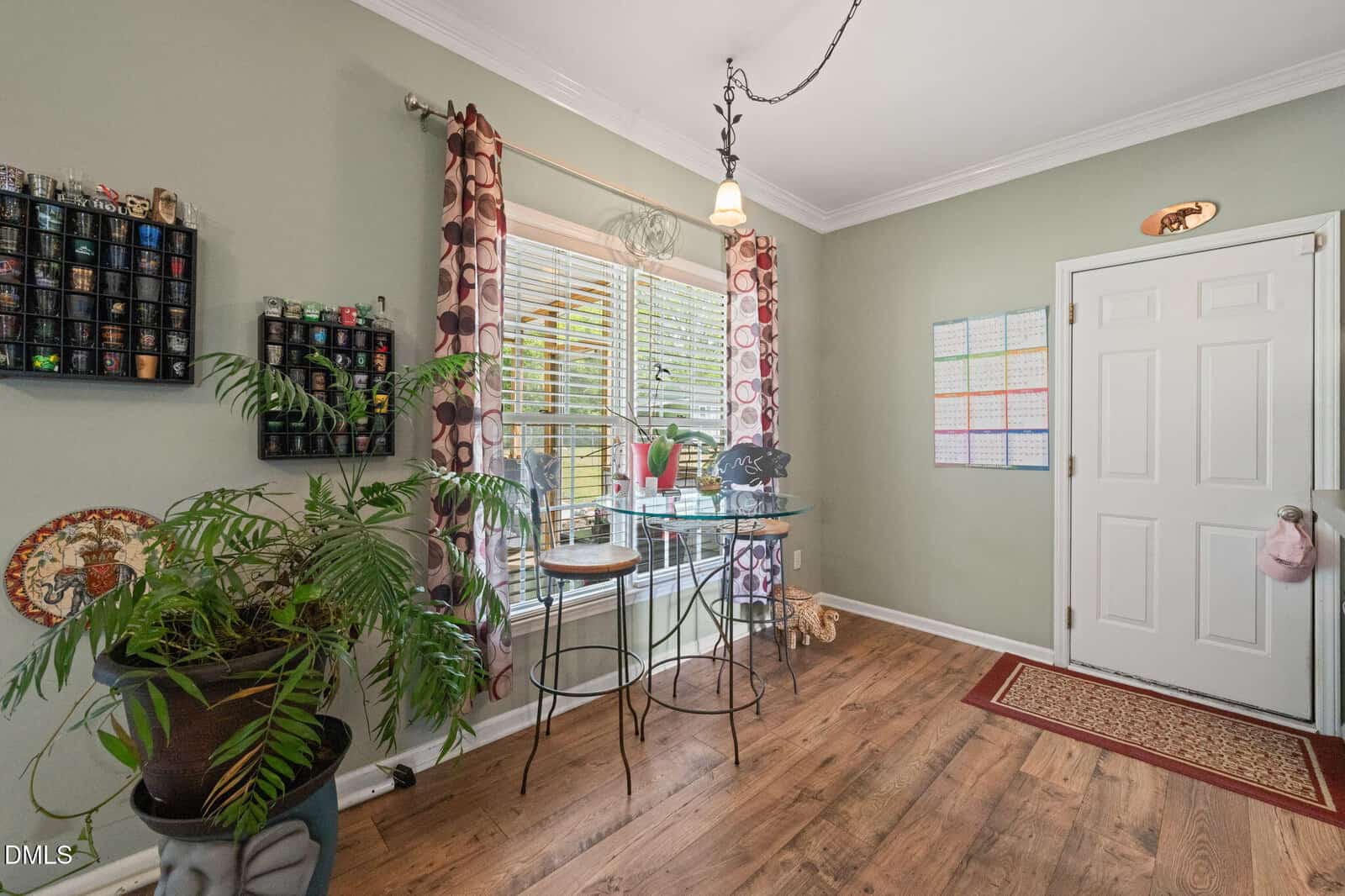 A bright room with wood flooring, green walls, a potted plant, display shelves, a table by a large window with patterned curtains, and a white door with a colorful calendar on the wall beside it.
