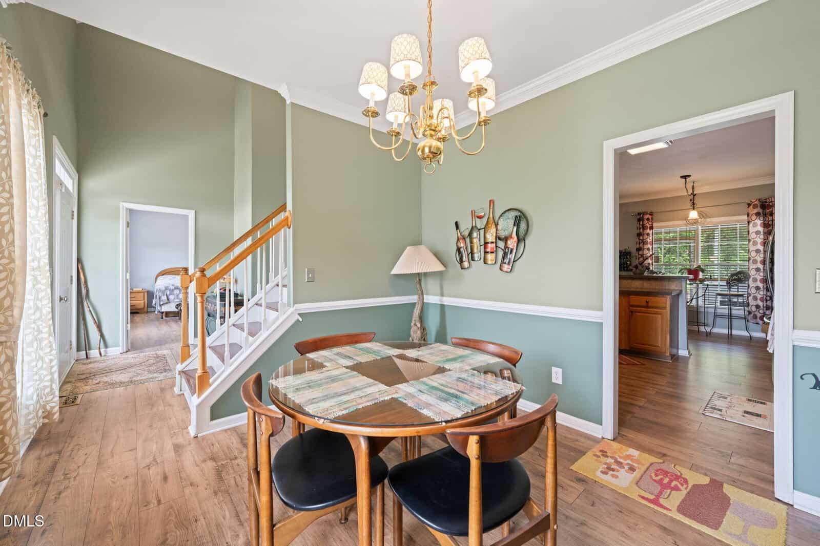 A dining area with a round table and four chairs, a chandelier above, sage green walls, wood floors, and a partial view of the kitchen, staircase, and adjacent room.