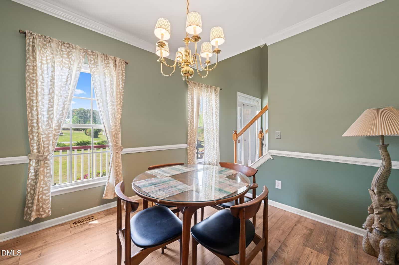 A dining area with a round table and four chairs, a chandelier overhead, green walls with white trim, a window with curtains, and a floor lamp shaped like an elephant. Stairs and front door are visible in the background.
