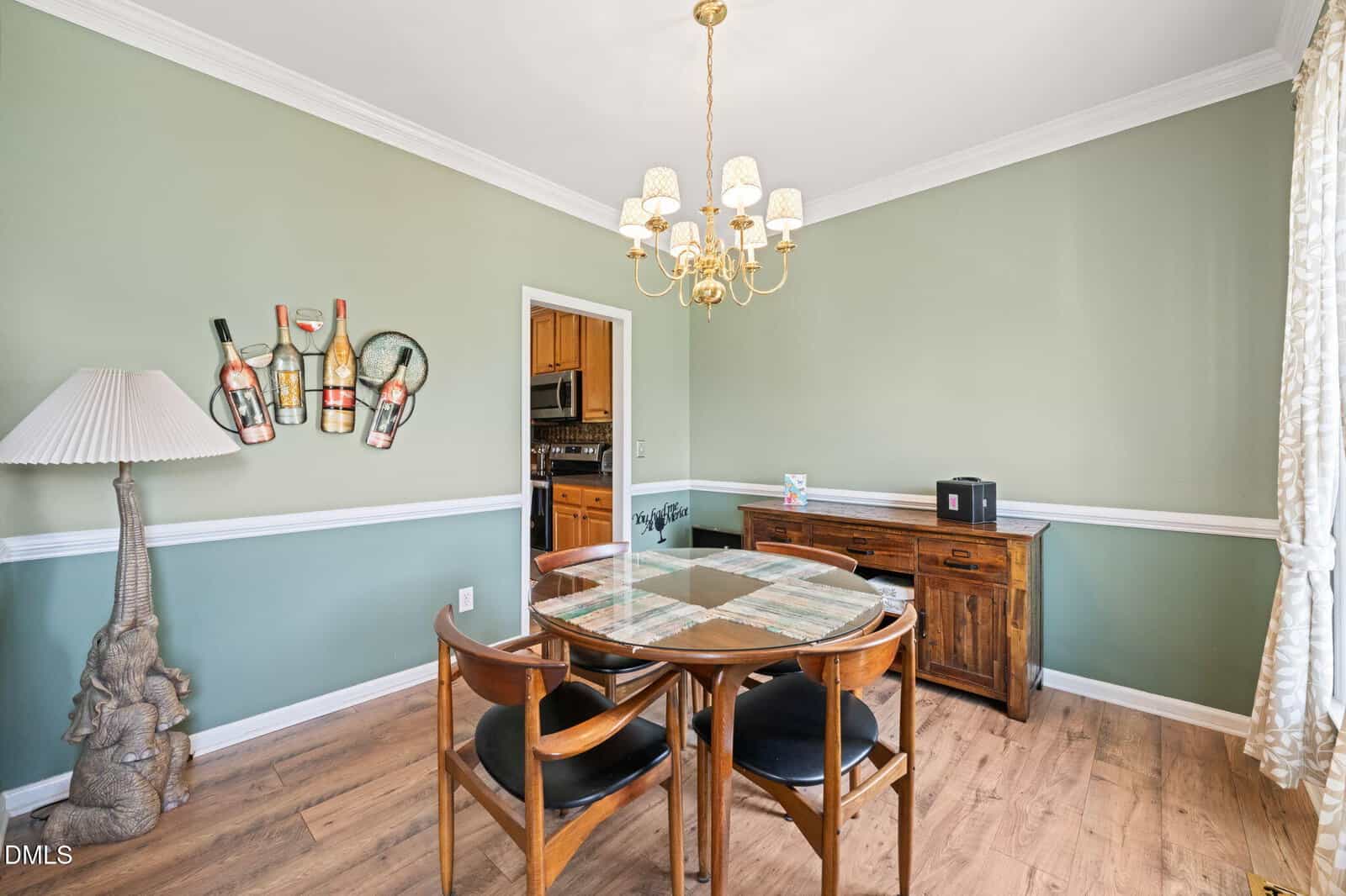Dining room with green walls, a chandelier, a wood table with four chairs, a sideboard, wall art of wine bottles, an elephant-shaped lamp, hardwood floors, and a window with curtain letting in natural light.