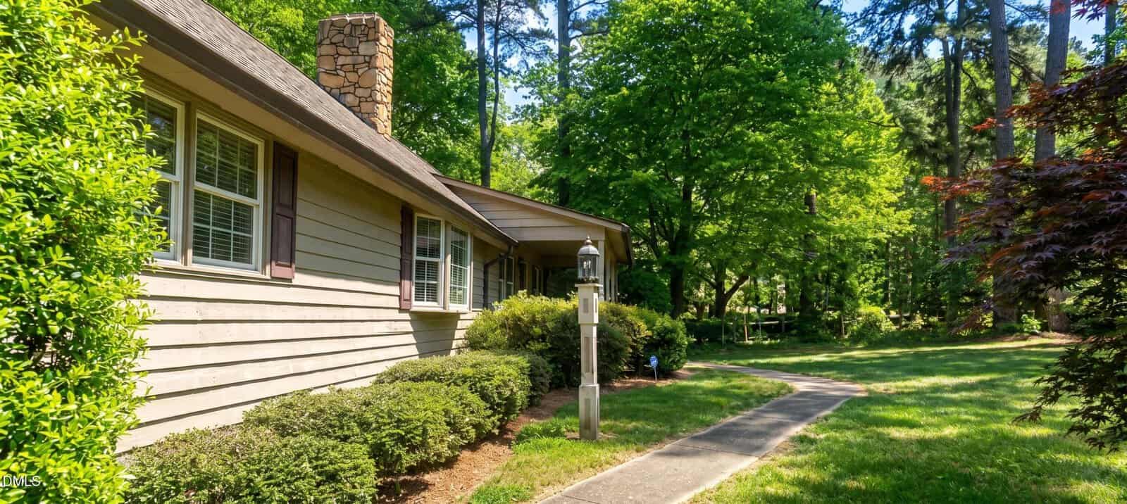 A beige house with white-trimmed windows, stone chimney, and covered porch is surrounded by lush green trees, bushes, and a trimmed lawn. A walkway leads to the entrance, passing a vintage-style lamp post.