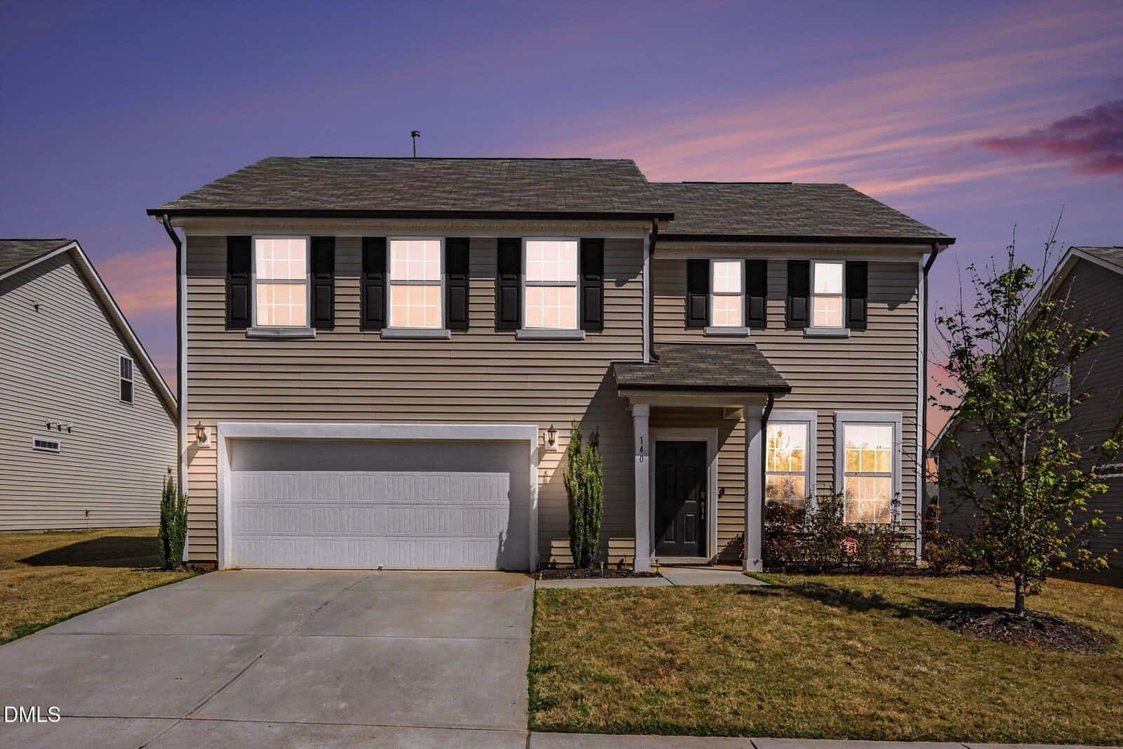 Two-story suburban house with beige siding, black shutters, a two-car garage, and a small front yard, photographed at sunset with a purple and pink sky in the background.
