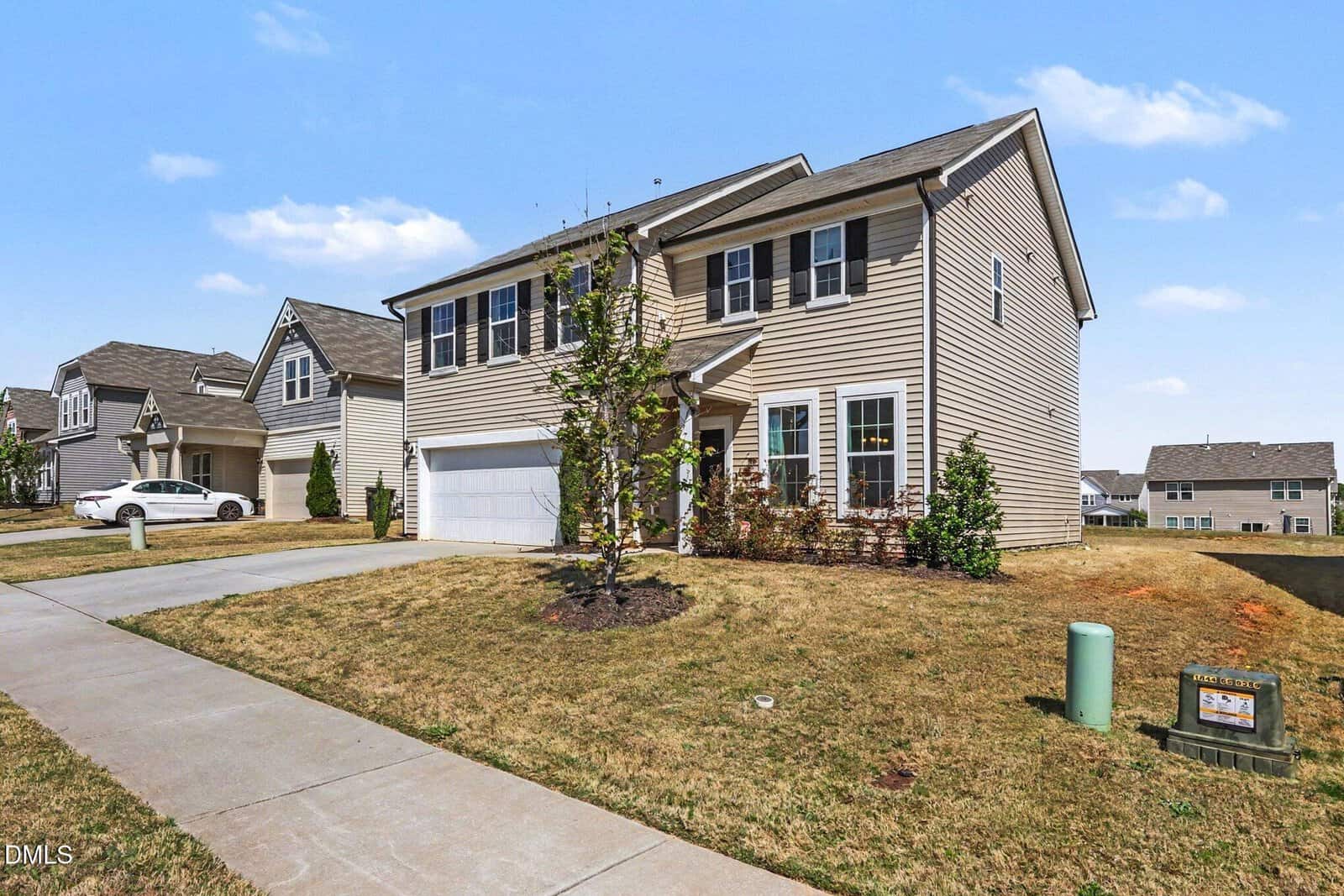 A two-story beige house with white trim and shutters, a double garage, and a sloping driveway, set on a grassy yard with young trees and shrubs, in a suburban neighborhood under a clear blue sky.