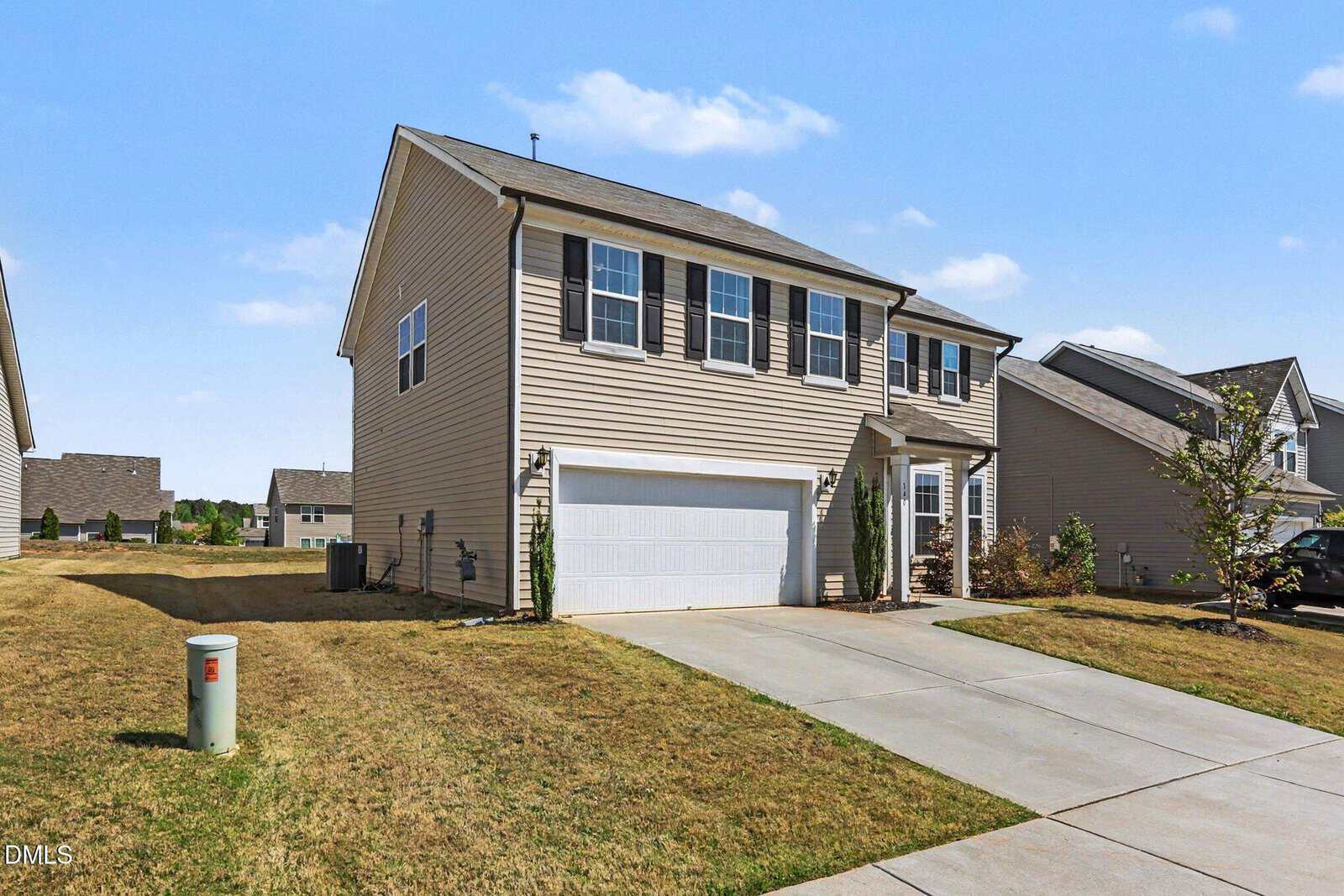 A two-story beige house with black shutters, a white garage door, and a concrete driveway, set on a neatly mowed lawn in a suburban neighborhood under a blue sky with scattered clouds.