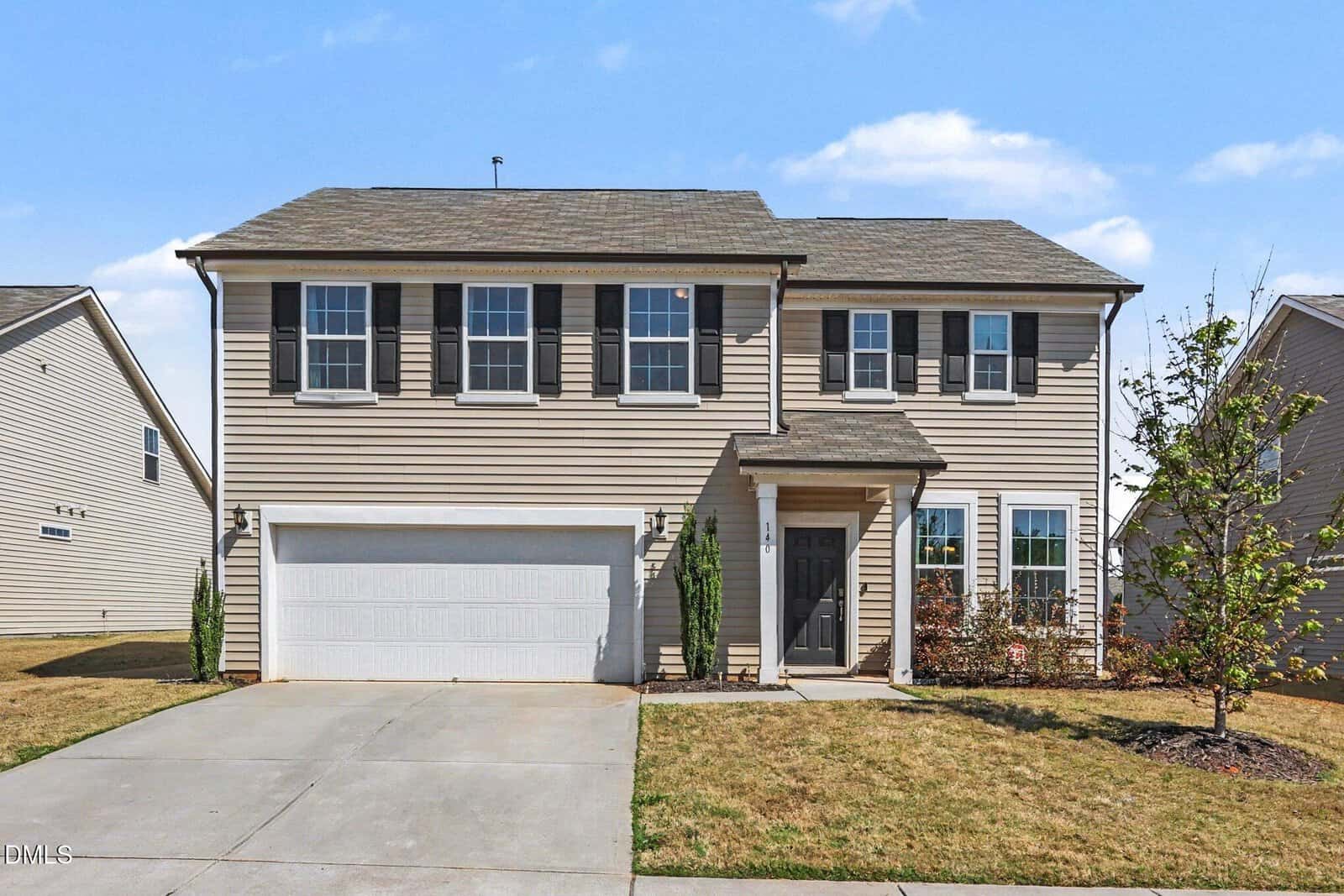A two-story suburban house with beige siding, black shutters, a two-car garage, a driveway, and a small front yard with young trees and shrubs under a blue sky.