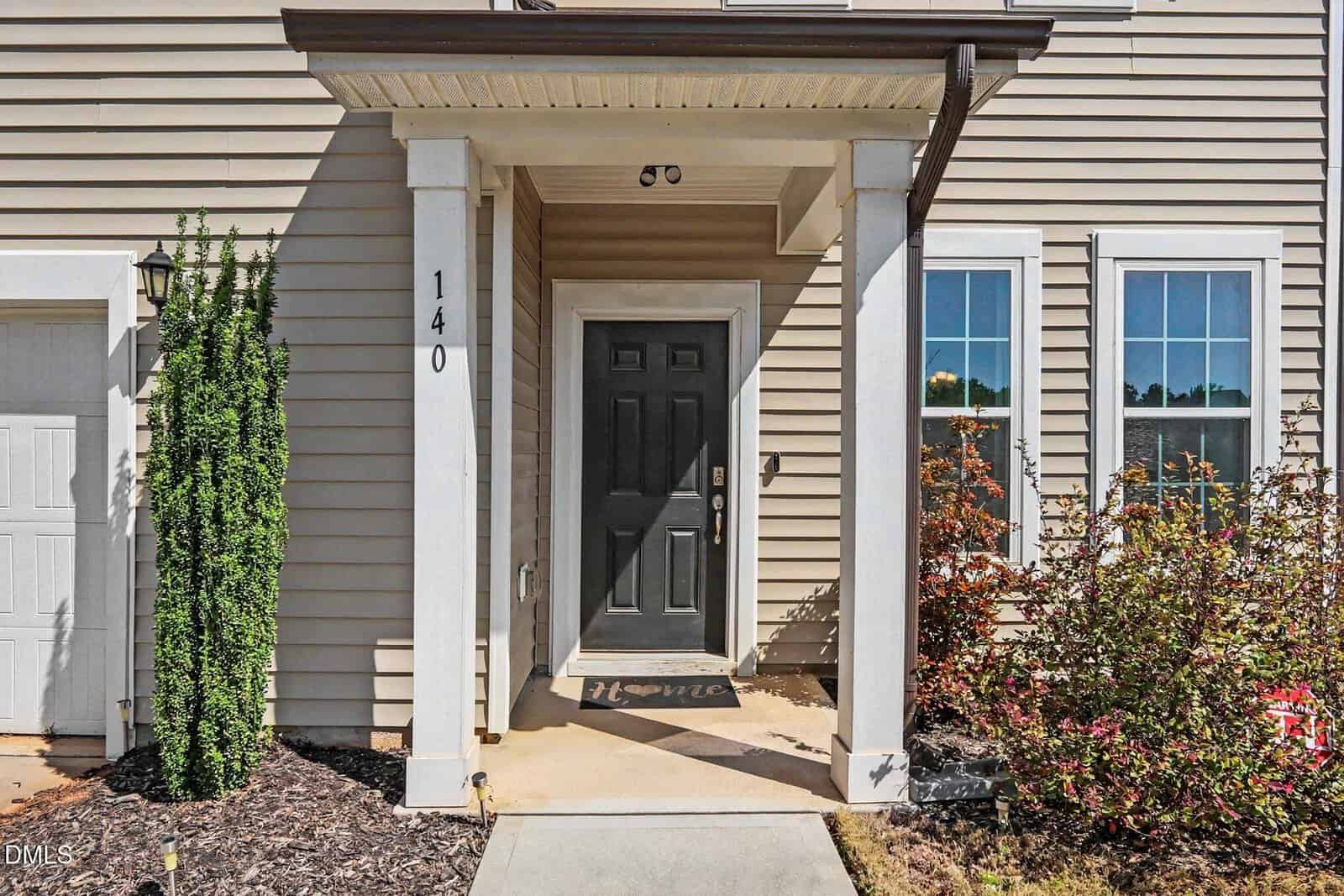 A tan house with white trim, black front door, two white columns, and the number 140 by the entrance. Bushes and plants are on either side of the walkway leading to the porch.