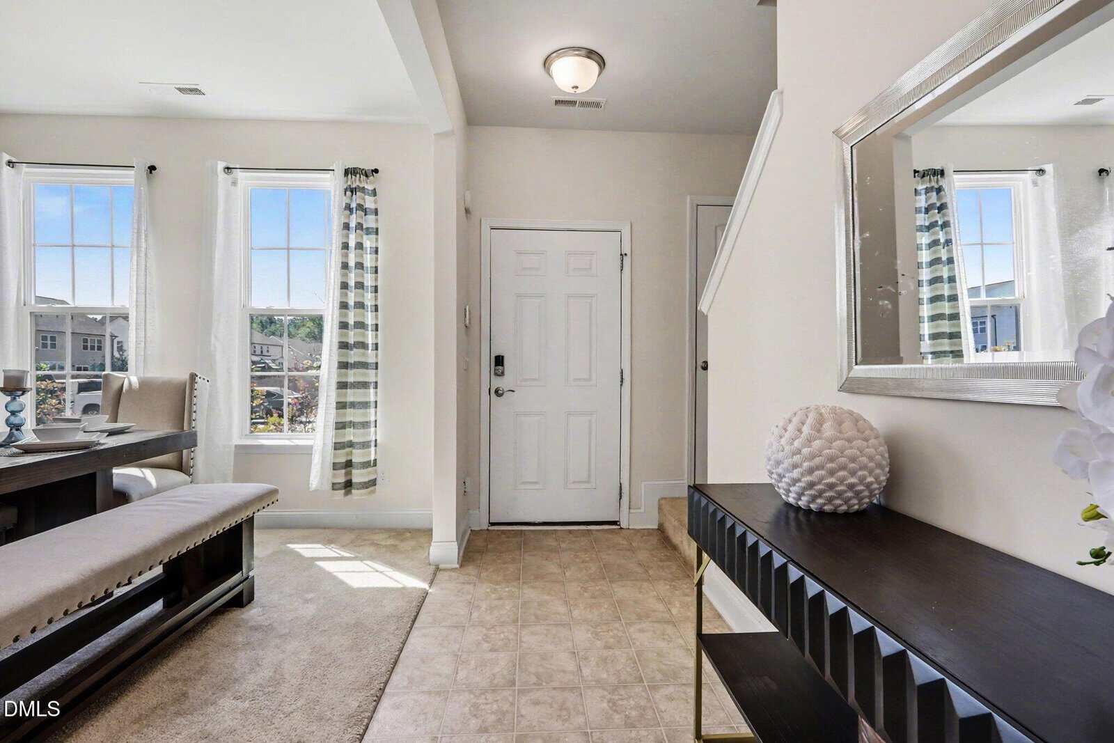 Bright entryway with a white front door, tile flooring, a black console table with decor, and a large wall mirror. Dining area with windows and striped curtains is visible on the left.