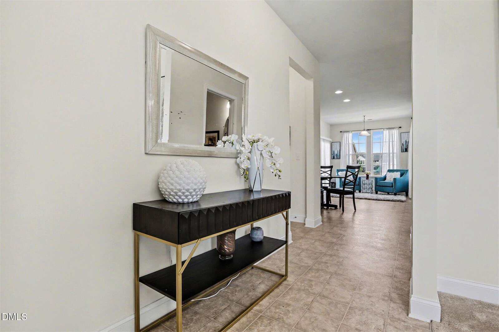 A hallway with a black console table, white vase, and flowers under a large mirror. The hallway leads to a dining area and a living room with blue accents, neutral walls, and tiled floors.