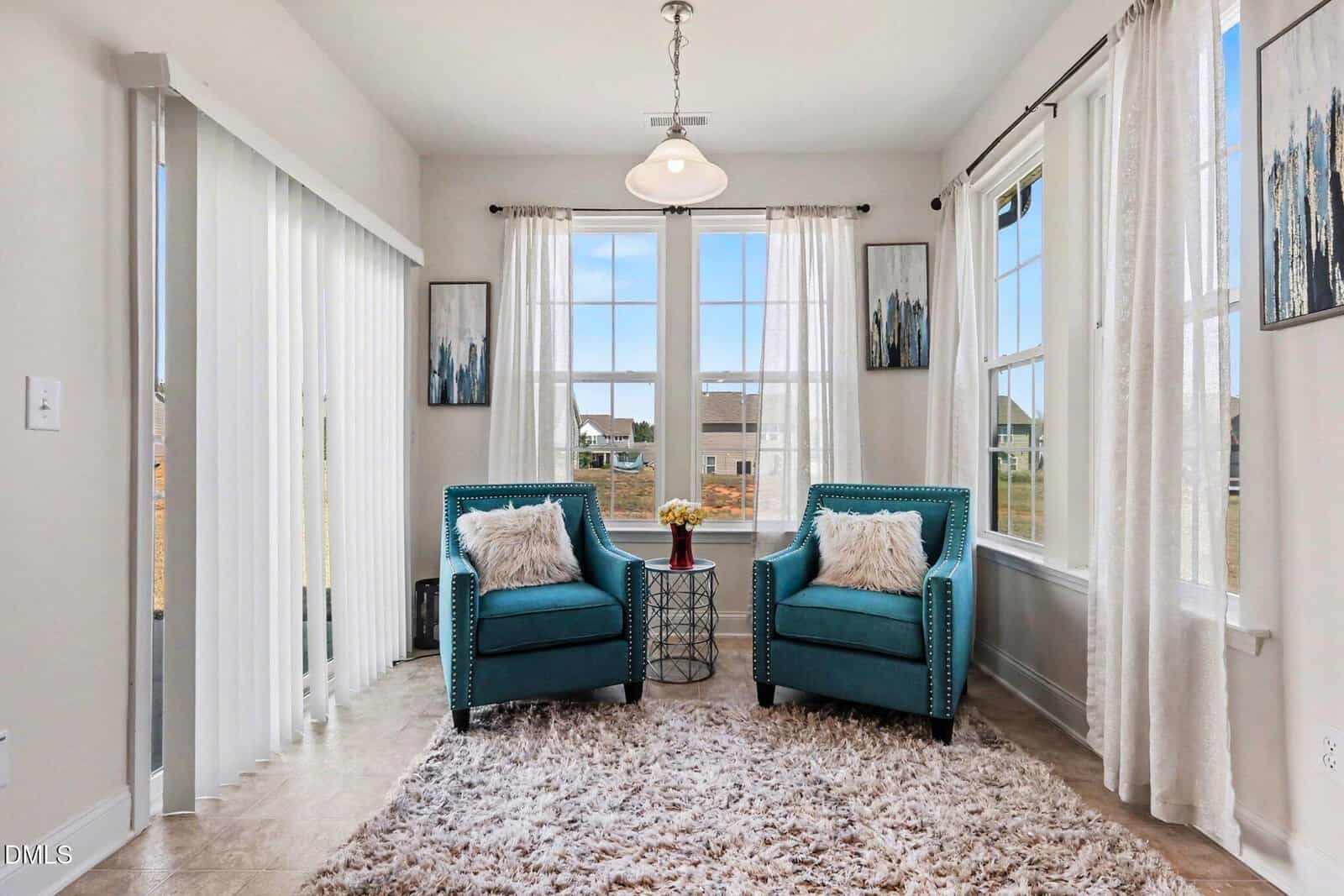 Bright sitting area with two teal armchairs, a round side table, and a fluffy rug. Large windows with white curtains let in natural light, and modern artwork decorates the walls.