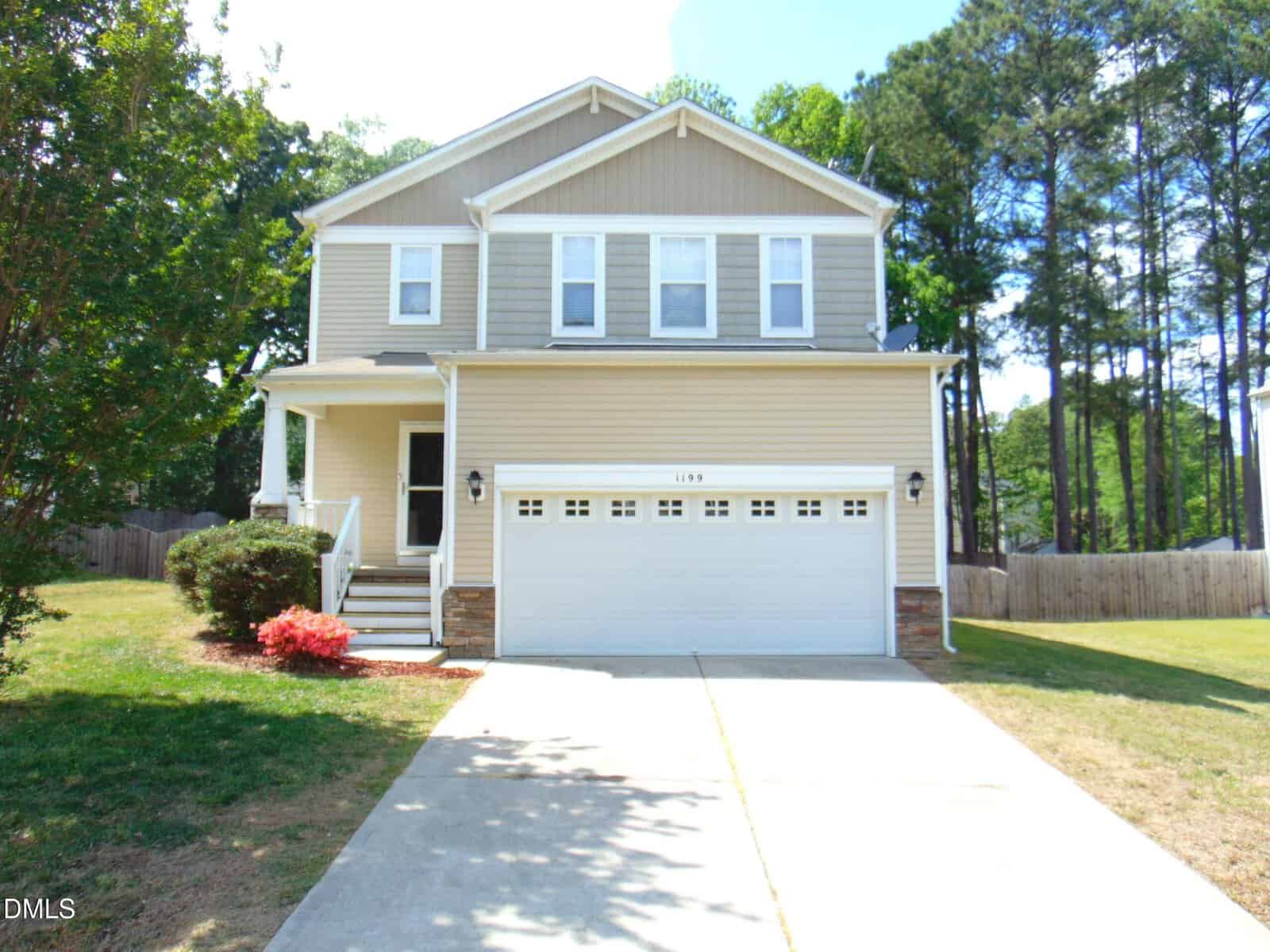 Two-story beige house with white trim, a front porch, and a two-car garage. The house has a large driveway, green lawn, and is surrounded by trees and a wooden fence.