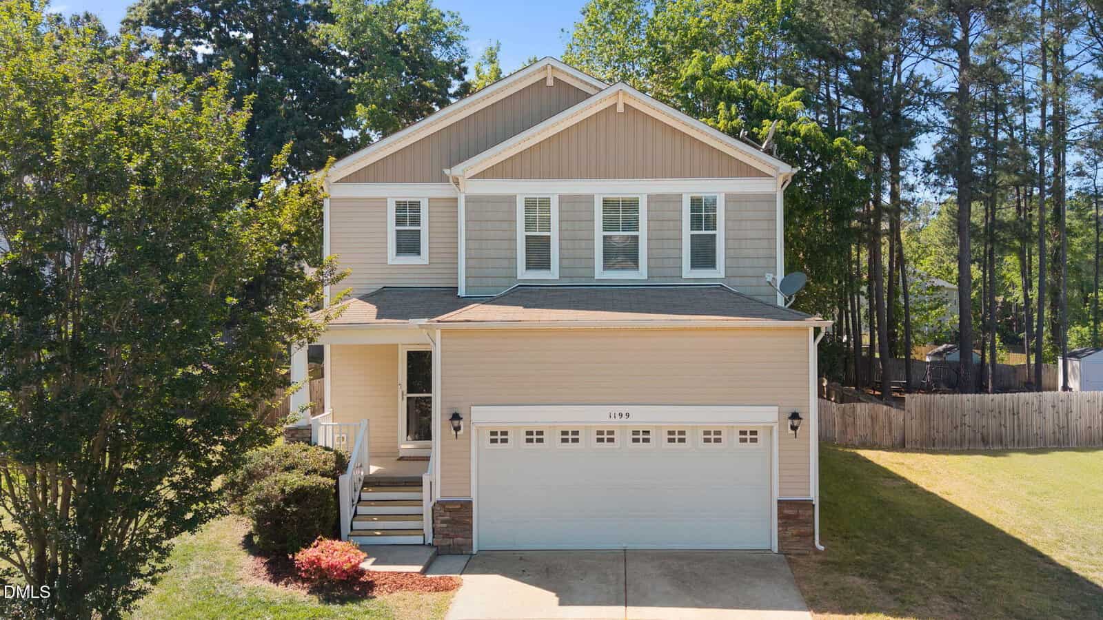 Two-story beige house with white trim and a double garage, surrounded by trees and a neatly maintained lawn. Steps lead to a small front porch, and a wooden fence runs along the right side of the backyard.