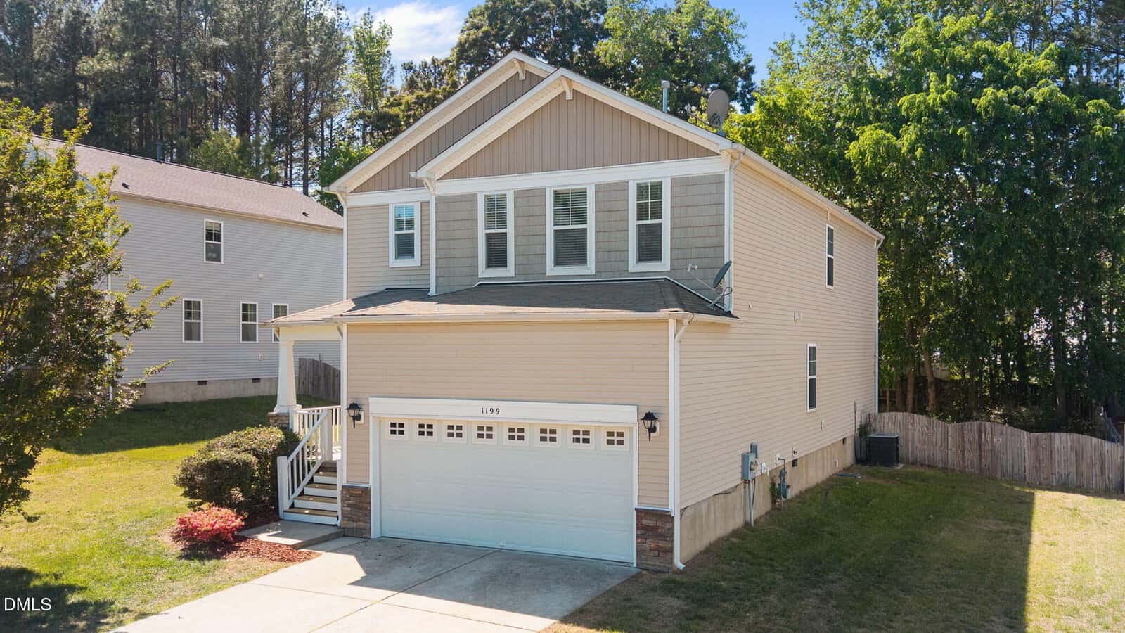 Two-story beige house with white trim, a double garage, front steps, and a small front yard. The house has multiple windows and is surrounded by trees and neighboring homes under a blue sky.