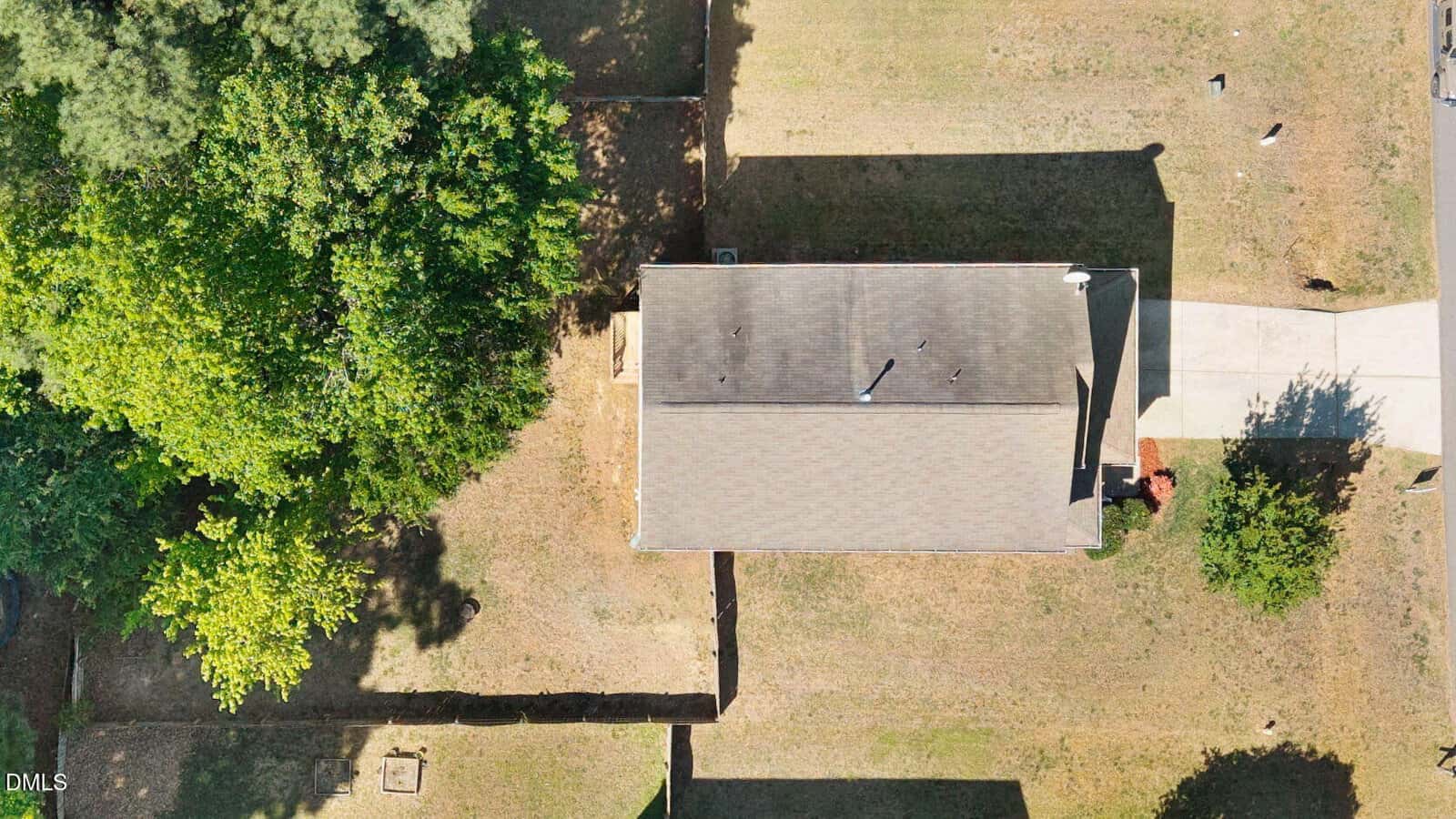 Aerial view of a single-story house with a gray roof, surrounded by a lawn, driveway, trees on the left, and patches of shade and sunlight. The property is bordered by fences and a sidewalk.
