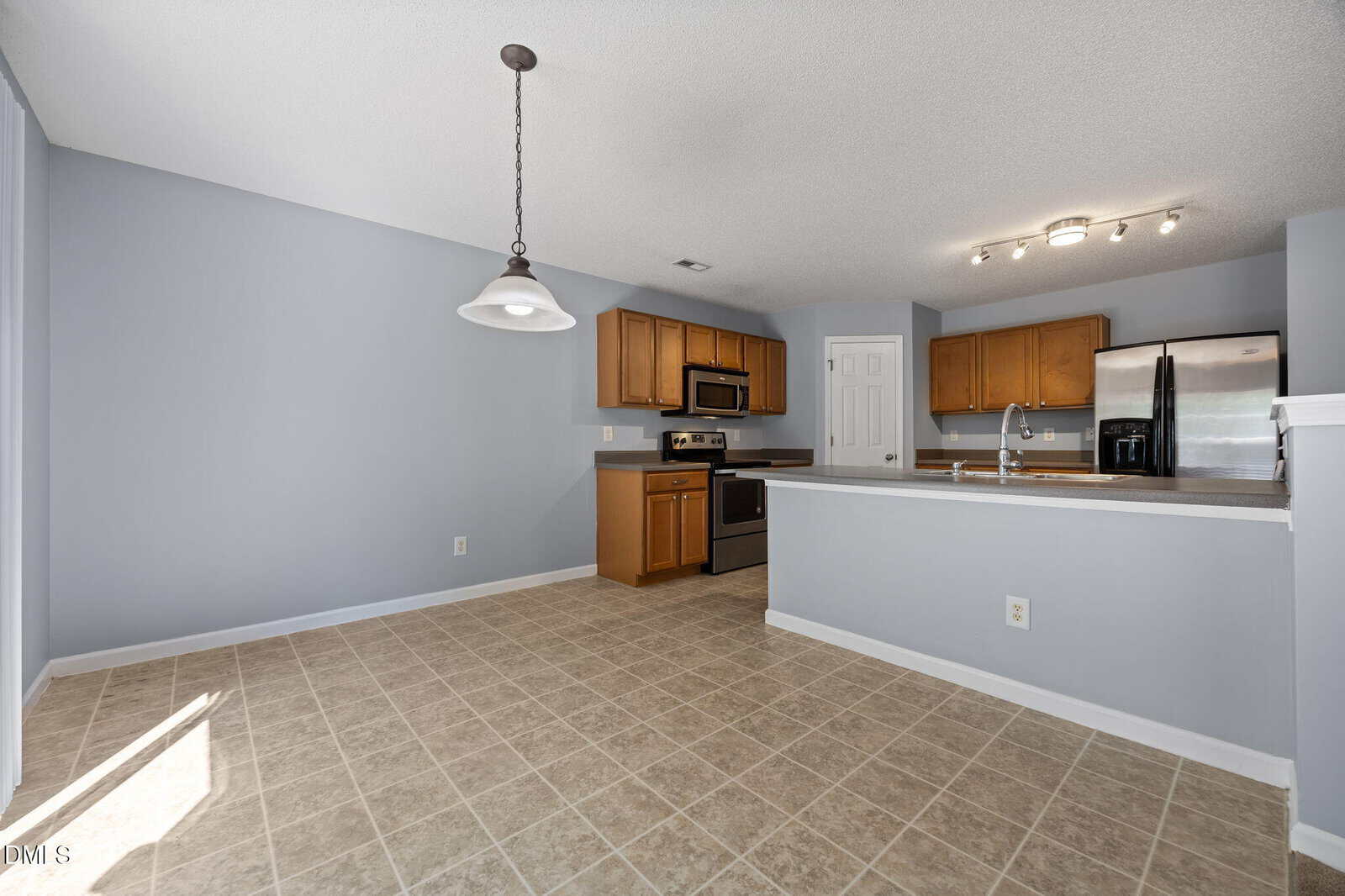 A spacious kitchen with light brown cabinets, stainless steel appliances, gray countertops, and an adjoining dining area with tile flooring. A hanging light fixture is above the dining space. Walls are painted light blue.