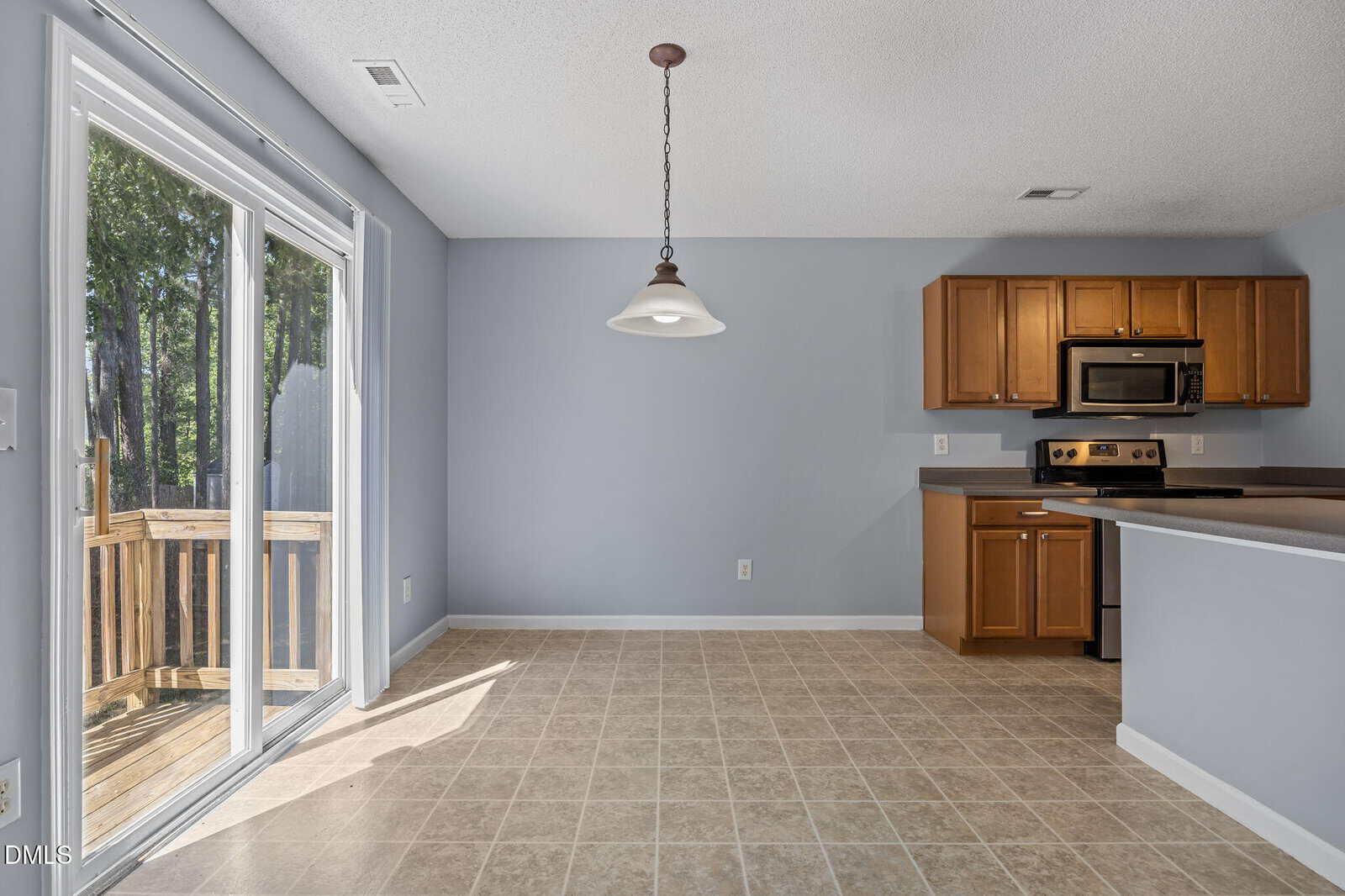 Kitchen with light brown cabinets, stainless steel appliances, tile floor, blue-gray walls, and a hanging light fixture. Large sliding glass door opens to a wooden deck with trees visible outside.