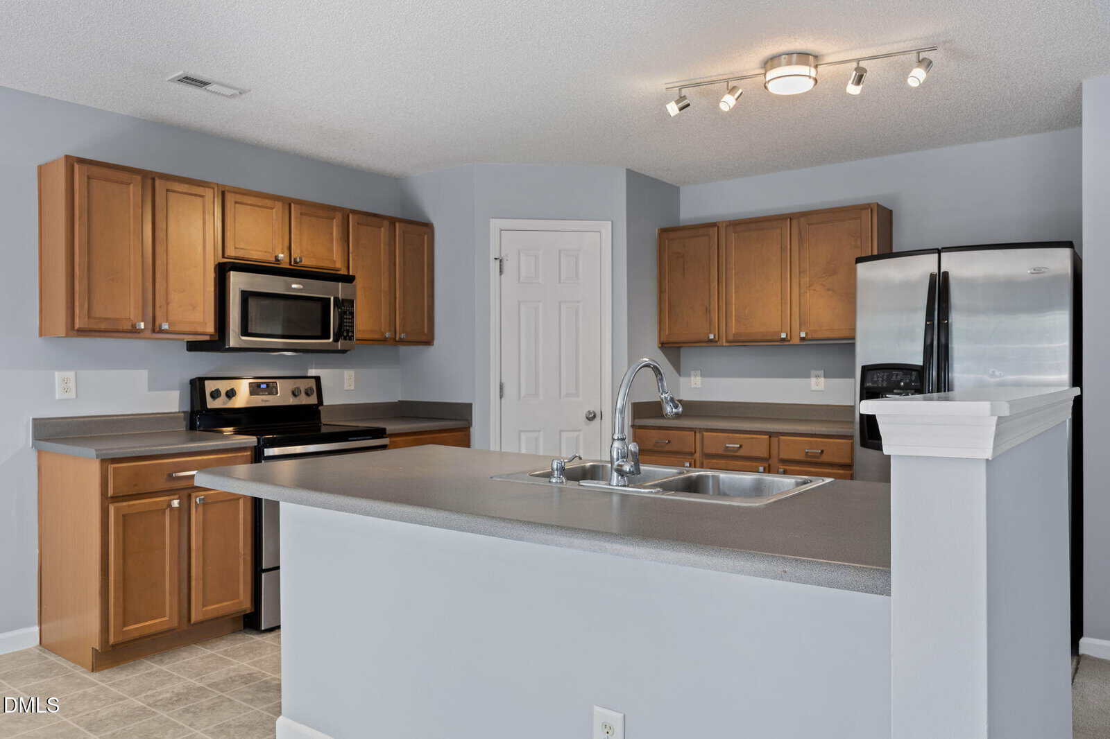 Modern kitchen with light brown cabinets, stainless steel appliances, a double sink on a gray countertop island, tiled flooring, and neutral gray walls with track lighting on the ceiling.