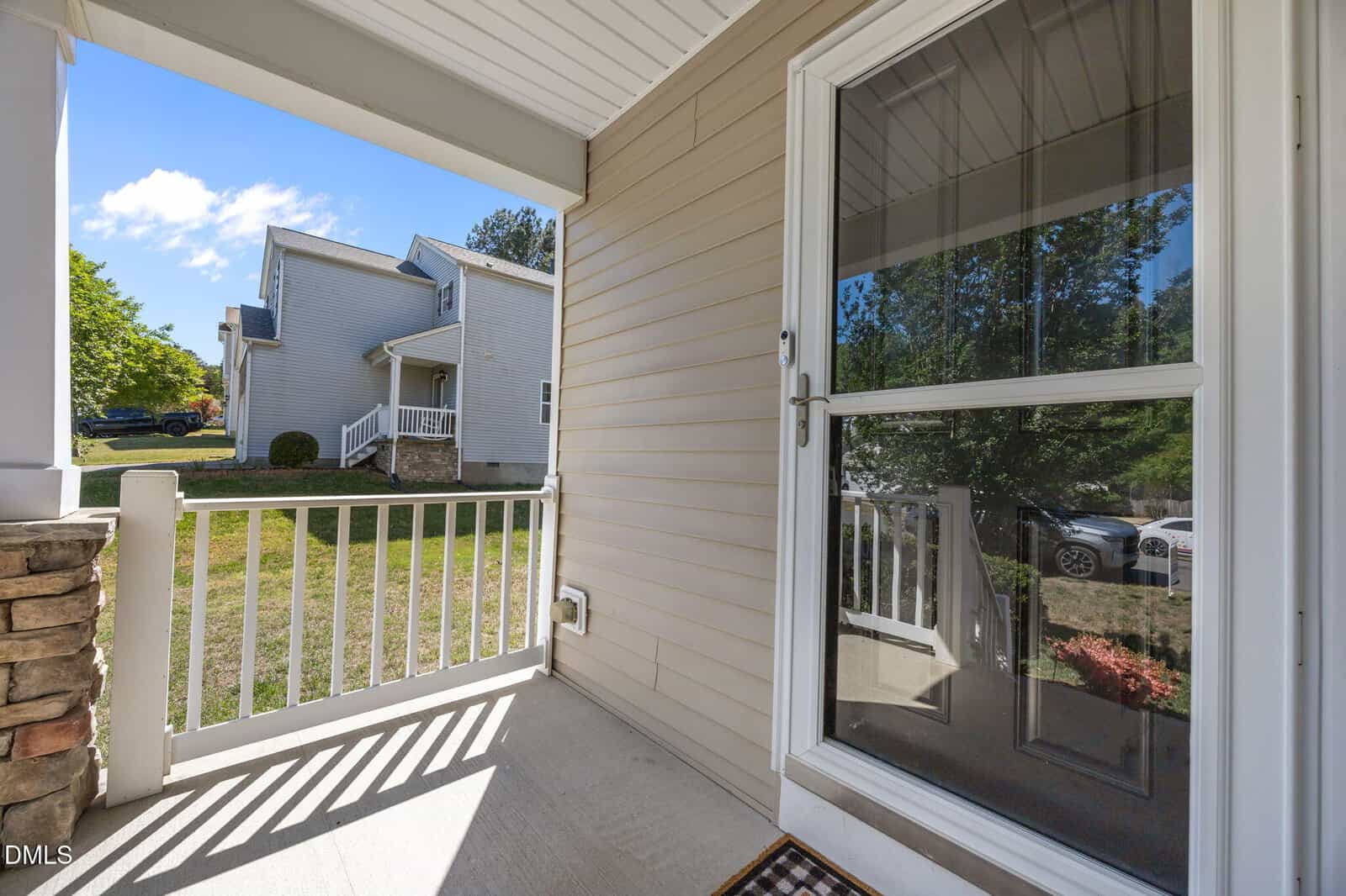 View from a covered front porch with beige siding and white railing, looking out toward a grassy yard and neighboring house under a blue sky with scattered clouds.
