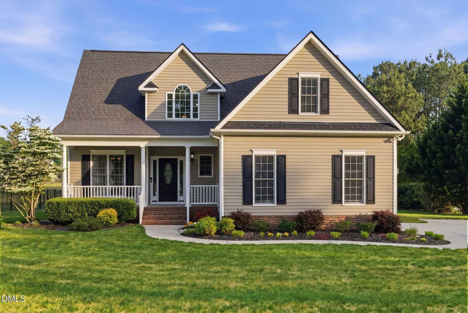 A two-story beige house with dark shutters, a covered front porch, and a well-manicured lawn with shrubs and flowers, set against a backdrop of trees under a blue sky.