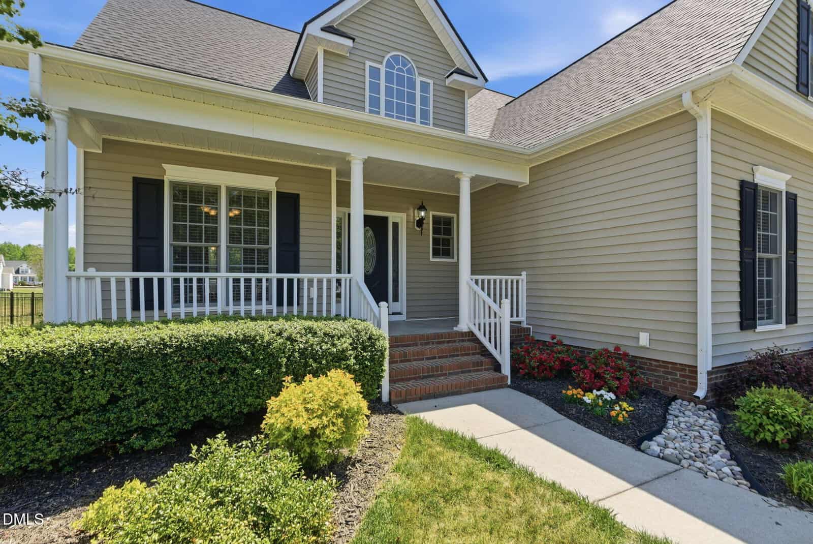 A beige house with white trim, black shutters, a covered front porch with railings, brick steps, a landscaped yard, and a curved concrete walkway leading to the front door.