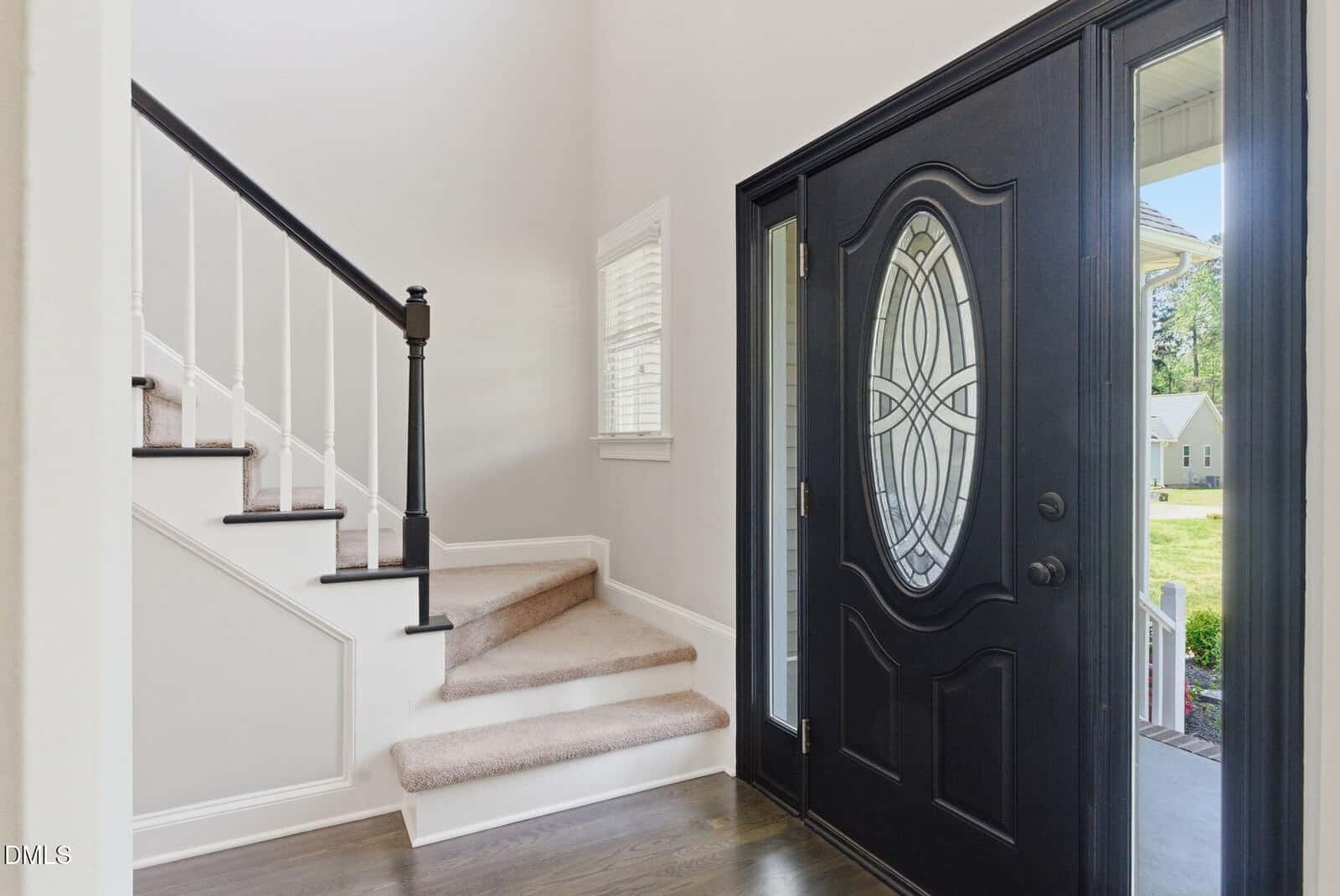 A front entryway with a black door featuring an oval glass window, and a staircase with white risers, light carpet, and a dark handrail. Sunlight streams through a window by the stairs.