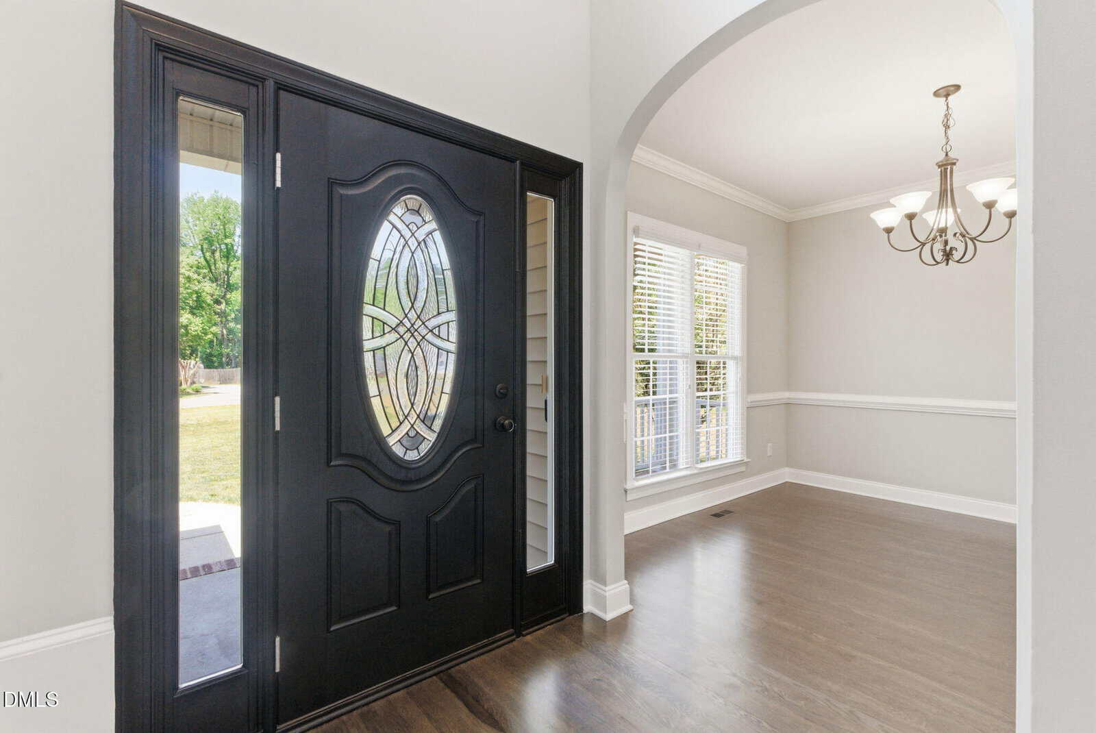 A black front door with decorative glass opens into a bright room with large windows, wood flooring, white trim, and a chandelier hanging from the ceiling.