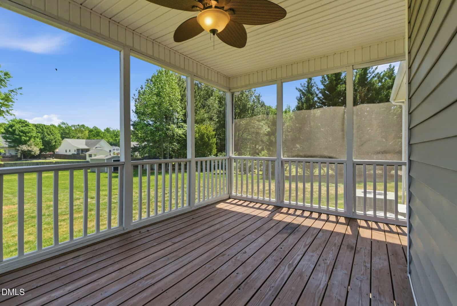 A screened-in porch with a ceiling fan and wooden floor overlooks a grassy backyard with trees and neighboring houses visible in the background.