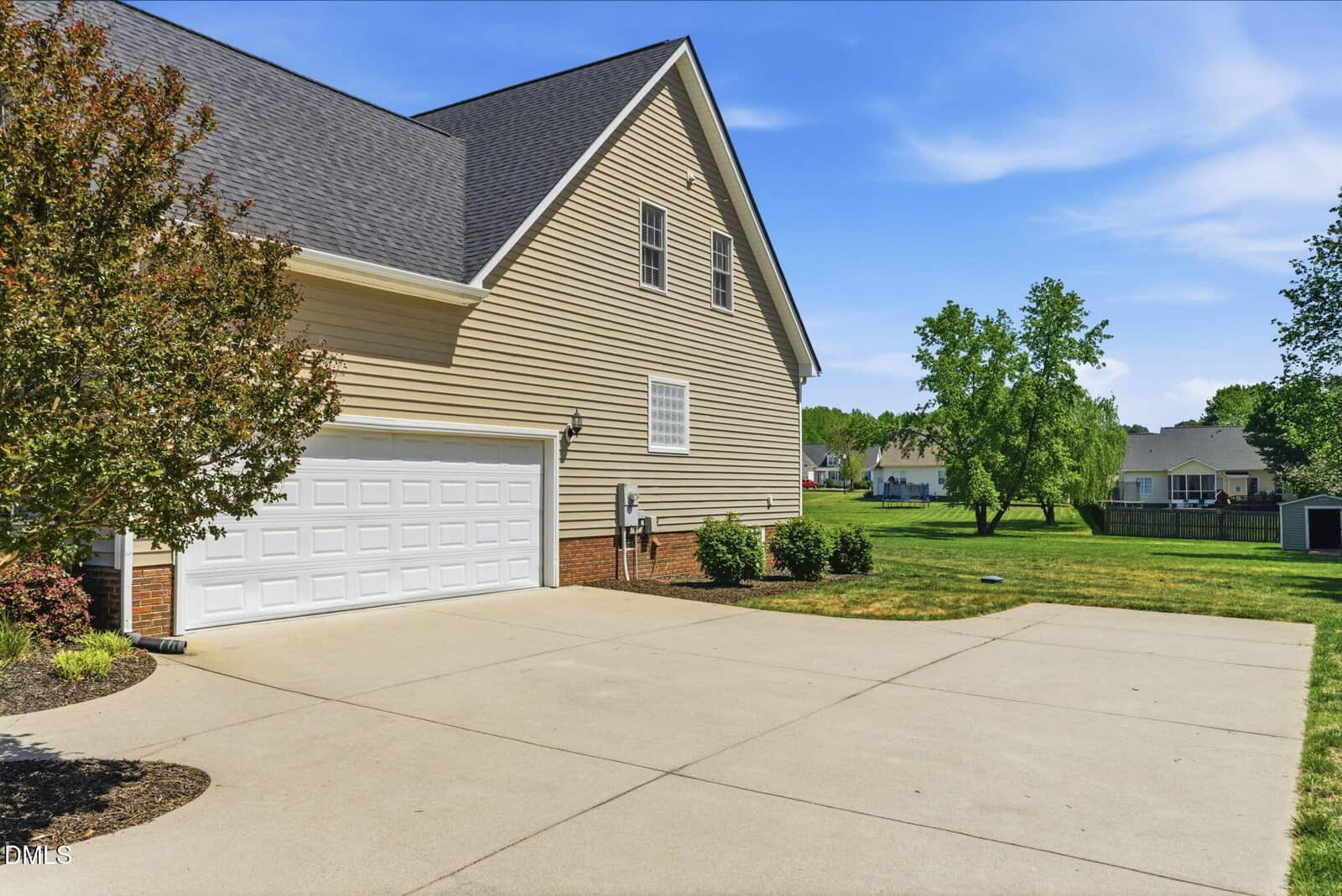 A beige house with a large white double garage door and a wide concrete driveway, surrounded by green lawn and trees under a clear blue sky.