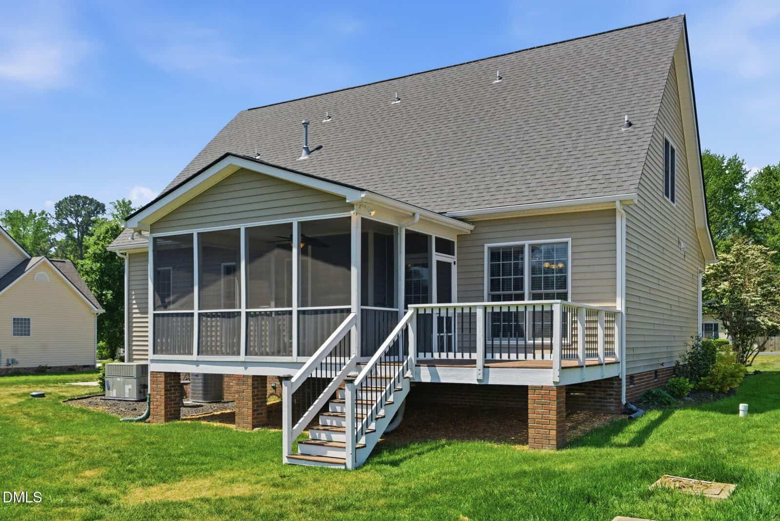 A beige house with a gray roof features a screened-in porch, wooden deck, and stairs leading to a grass lawn. The house sits on a brick foundation and is surrounded by trees and neighboring homes.