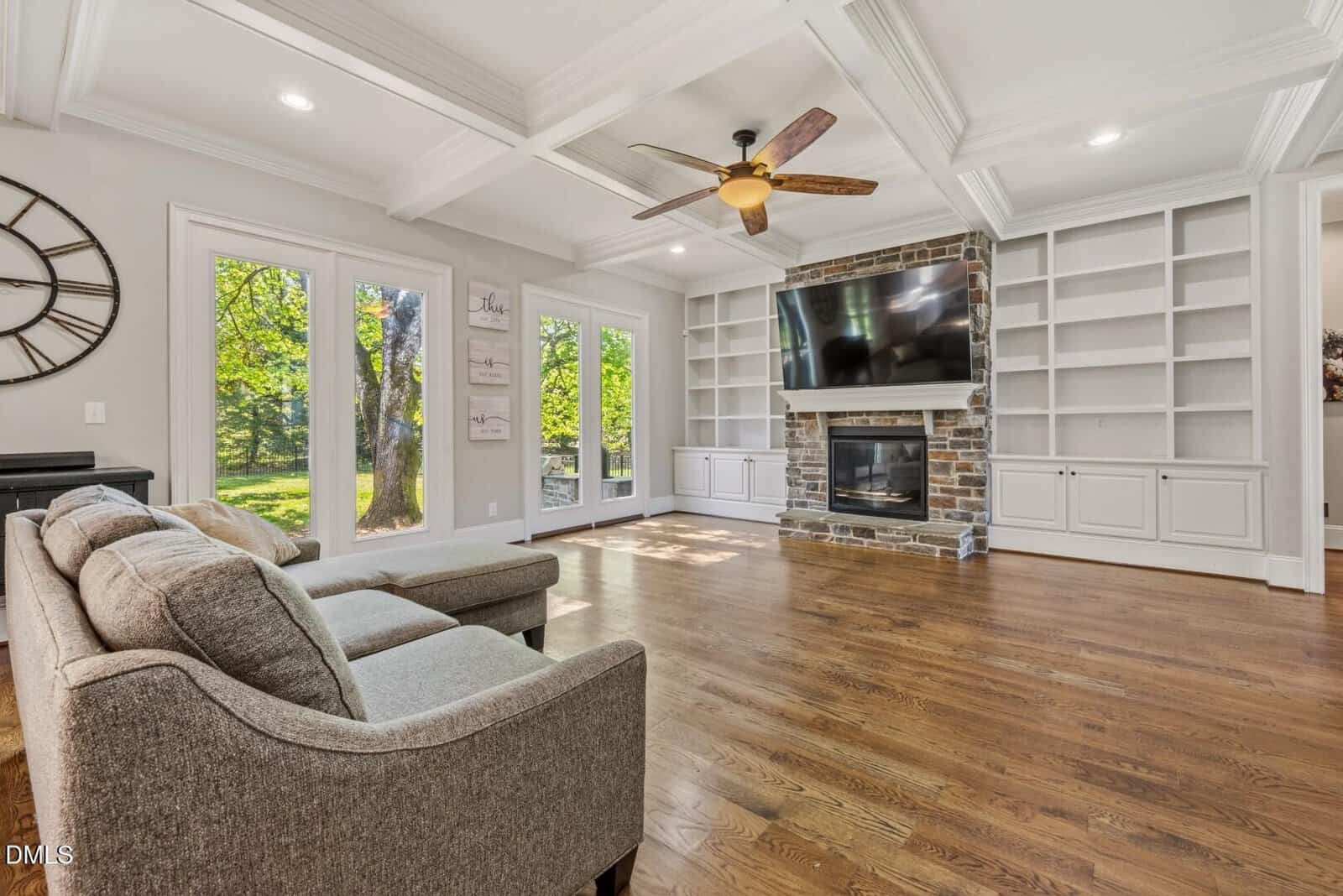 Bright living room with hardwood floors, gray sectional sofa, large windows, built-in shelves, stone fireplace with a mounted TV, ceiling fan, and coffered ceiling. Green trees are visible through the windows.