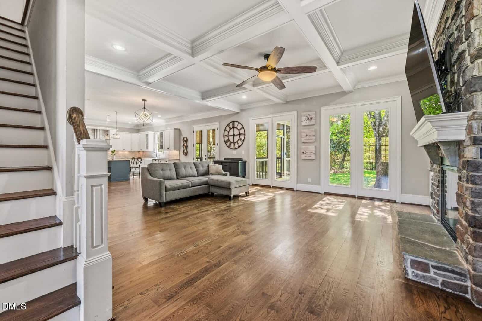 Spacious open-concept living room at 693 Hawthorne Place in Creedmoor features wooden floors, gray sectional sofa, coffered ceiling, stone fireplace, and large windows with French doors opening to a lush outdoor area. Kitchen visible in the background.