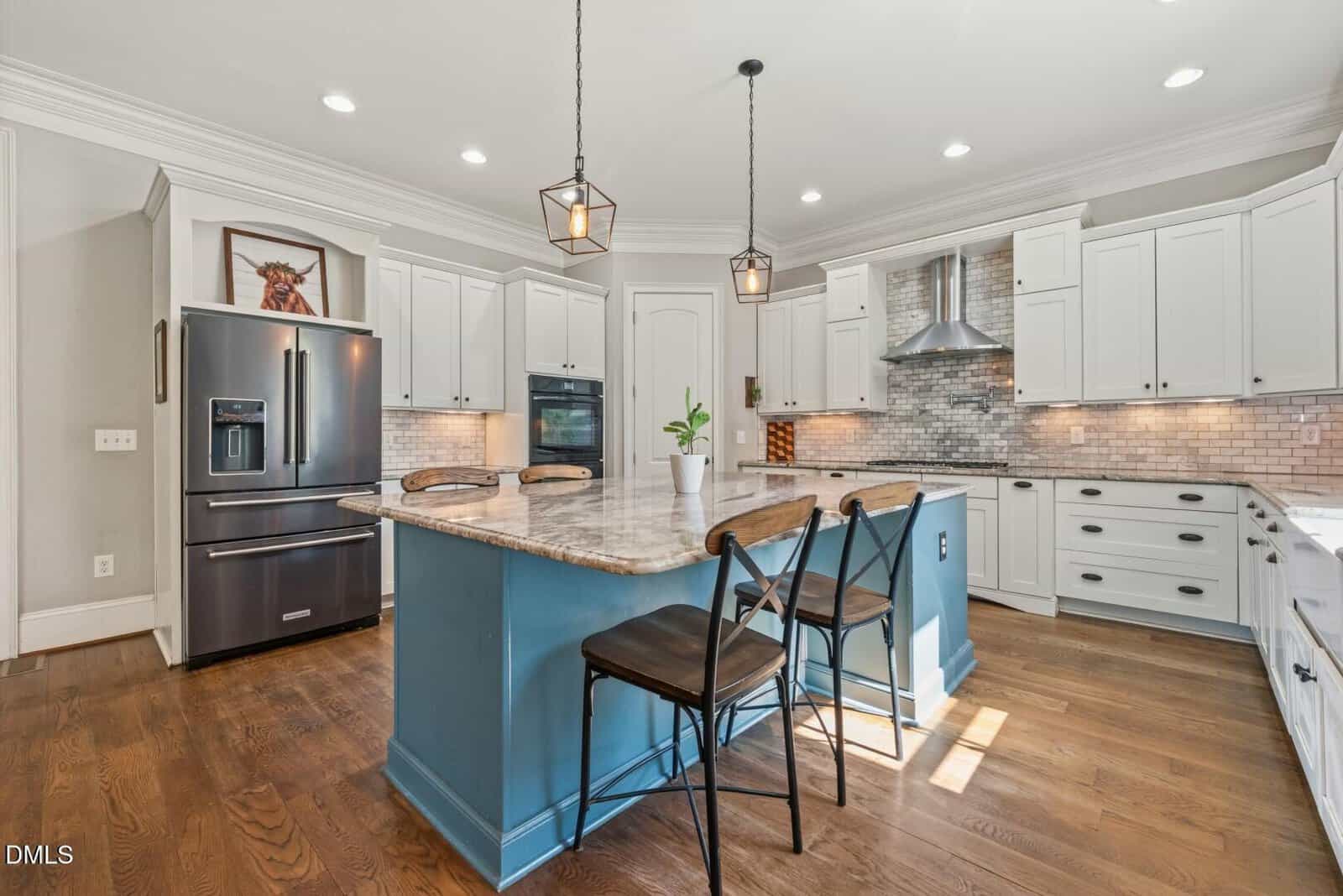 Modern kitchen with white cabinets, stainless steel appliances, a blue island with a marble countertop, two barstools, pendant lights above, and hardwood floors. There is a plant on the island and a horse artwork near the fridge.