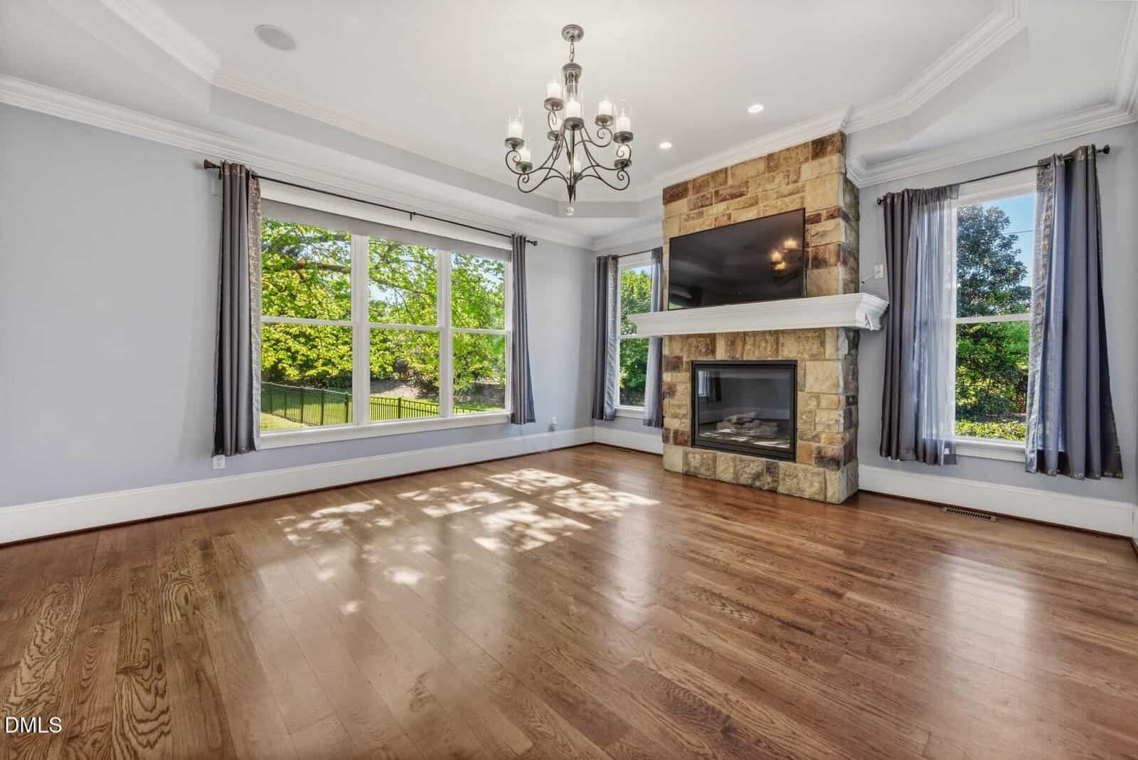 Bright living room with hardwood floors, large windows with gray curtains, a stone fireplace with a mounted TV, and a chandelier on the ceiling; lush greenery visible outside.