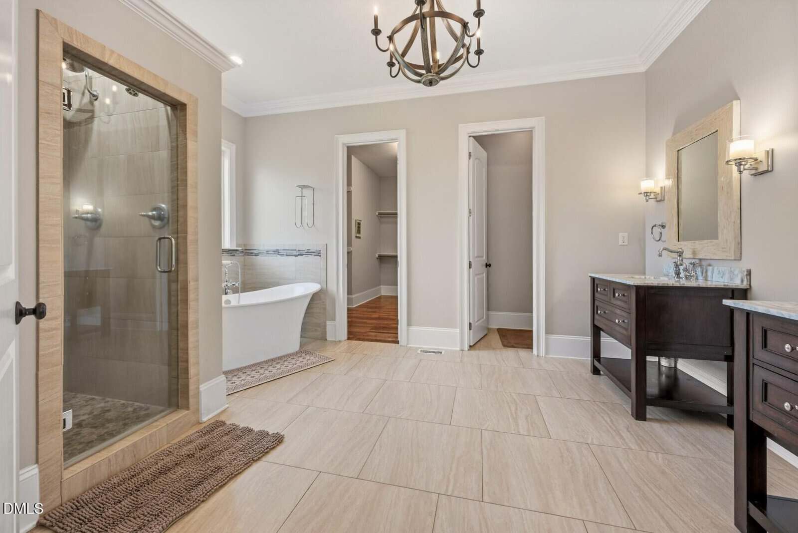 Spacious bathroom with a glass shower, freestanding tub, double sinks with dark wood vanities, large beige tile floor, chandelier, and neutral walls. Two doors lead to other rooms.