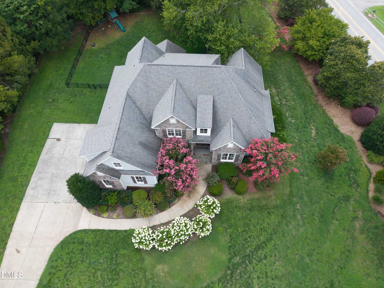 A two-story gray house with a large driveway and an attached garage, surrounded by green lawn, trees, and pink flowering shrubs, viewed from above. A road and playground are visible near the property.