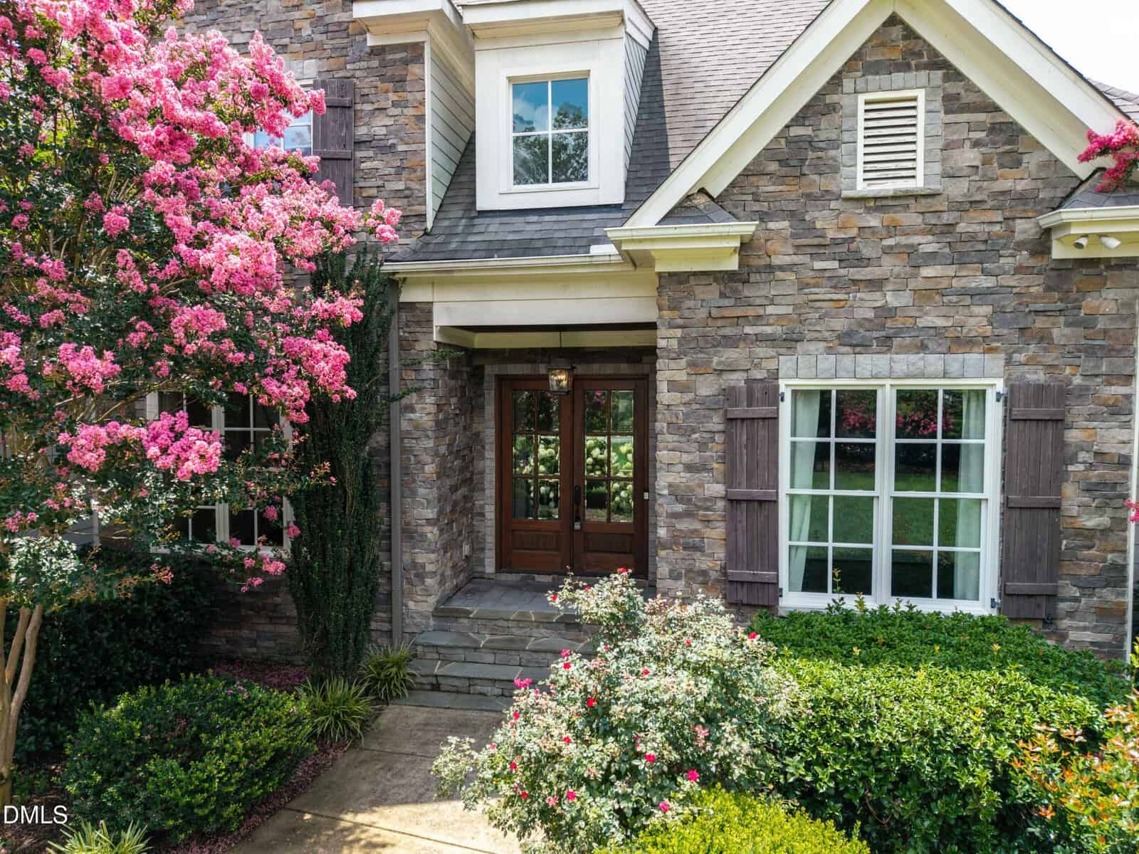 Front entrance of a stone house with double wooden doors, white trim, dormer window, and window shutters. Pink flowering trees and green bushes surround the stone walkway leading to the door.