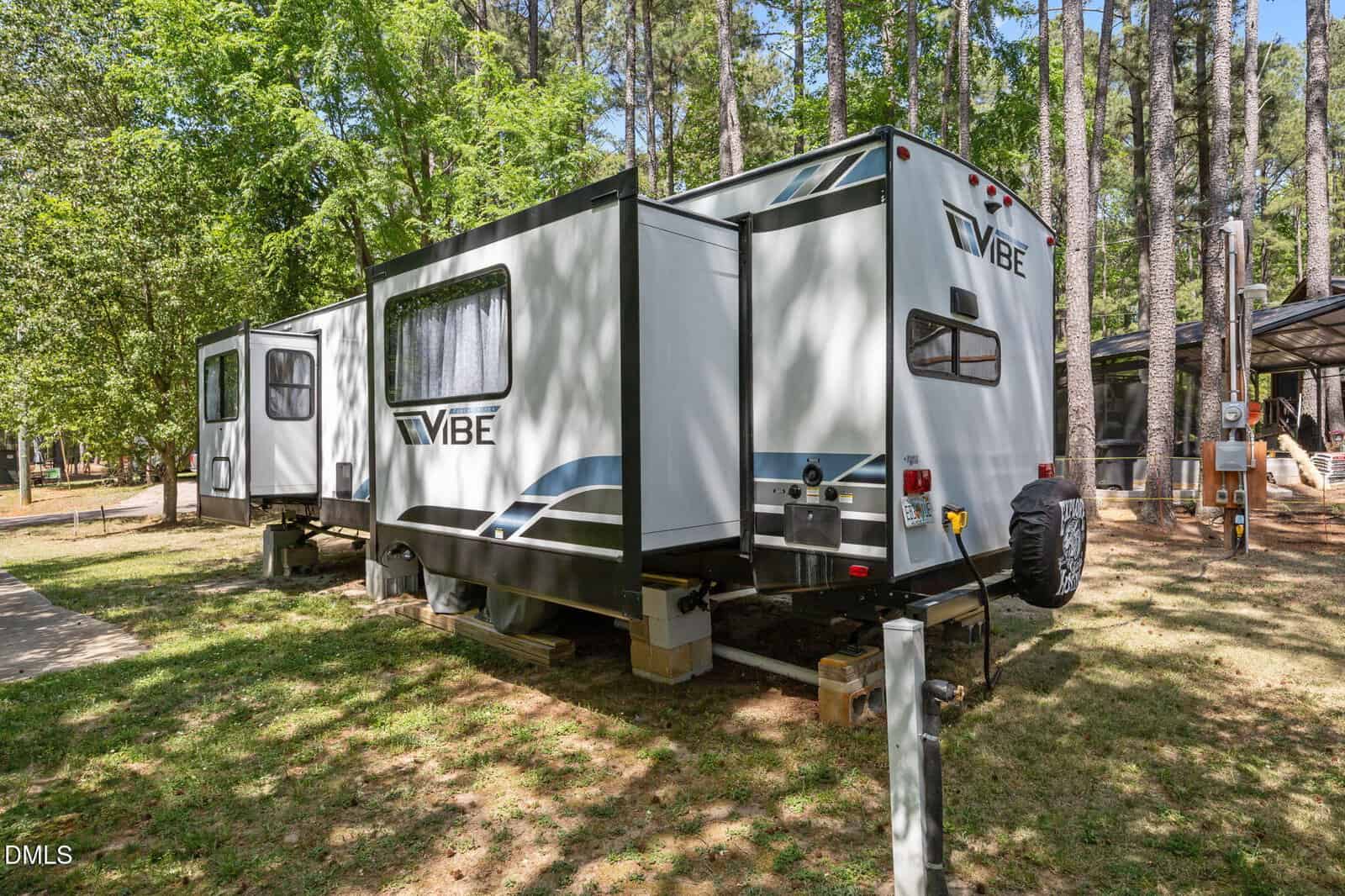 A white Vibe travel trailer with slide-outs is parked on a grassy lot among tall trees, elevated on blocks, with utility hookups visible nearby.